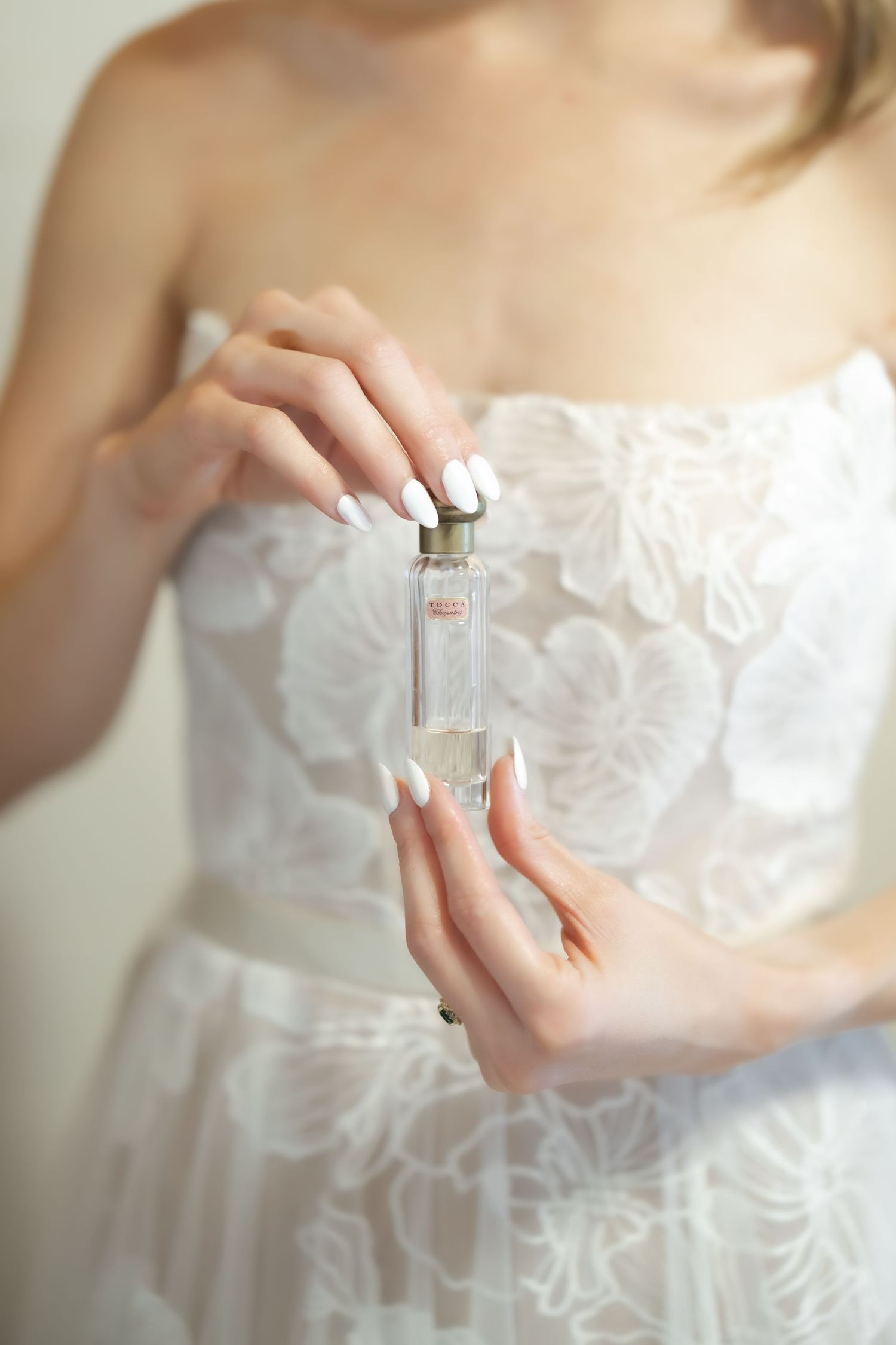 Bride in white lace dress holding a clear perfume bottle with both hands, white manicured nails.