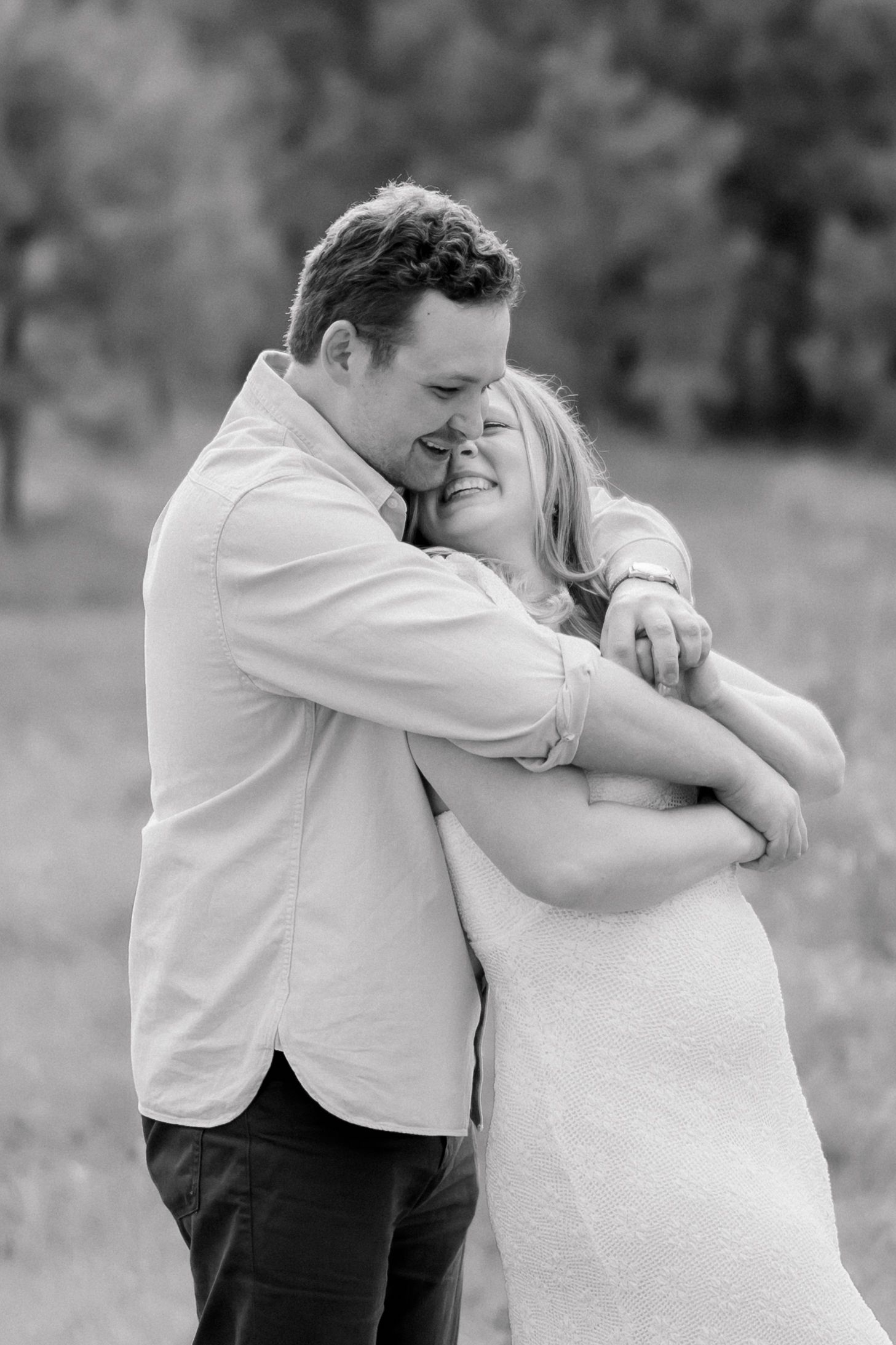 Black and white photo of man hugging woman from behind, both smiling joyfully outdoors.