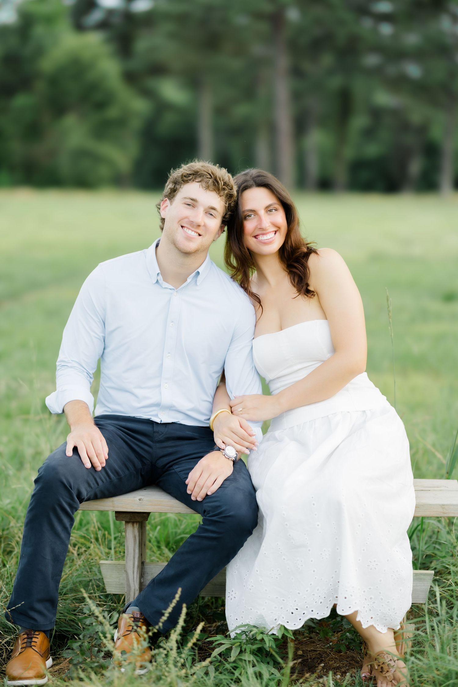 Smiling couple sitting on a wooden bench in a green field, woman in white eyelet dress leaning on mans shoulder.