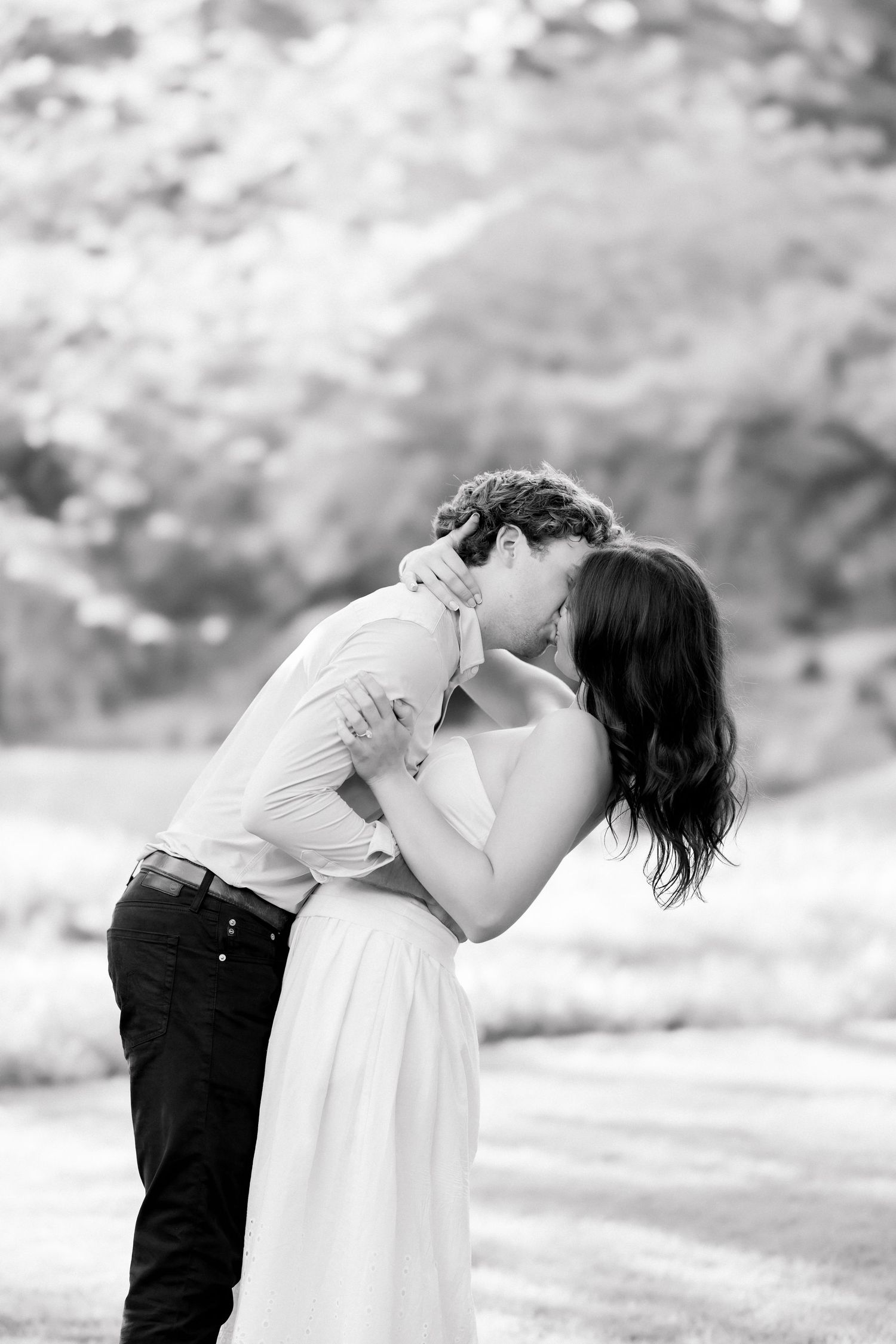 Black and white image of a couple sharing a tender kiss outdoors, woman in flowing white dress embracing her partner.