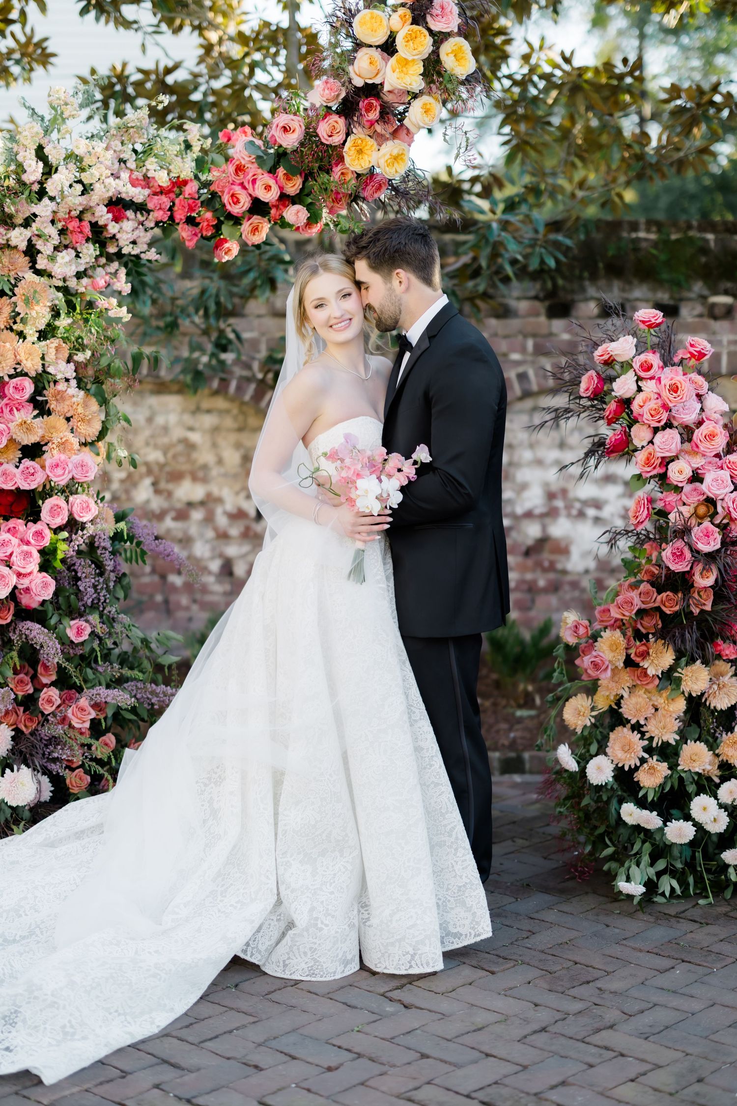A wedding couple poses under a stunning floral arch with pink and peach roses against a brick wall.