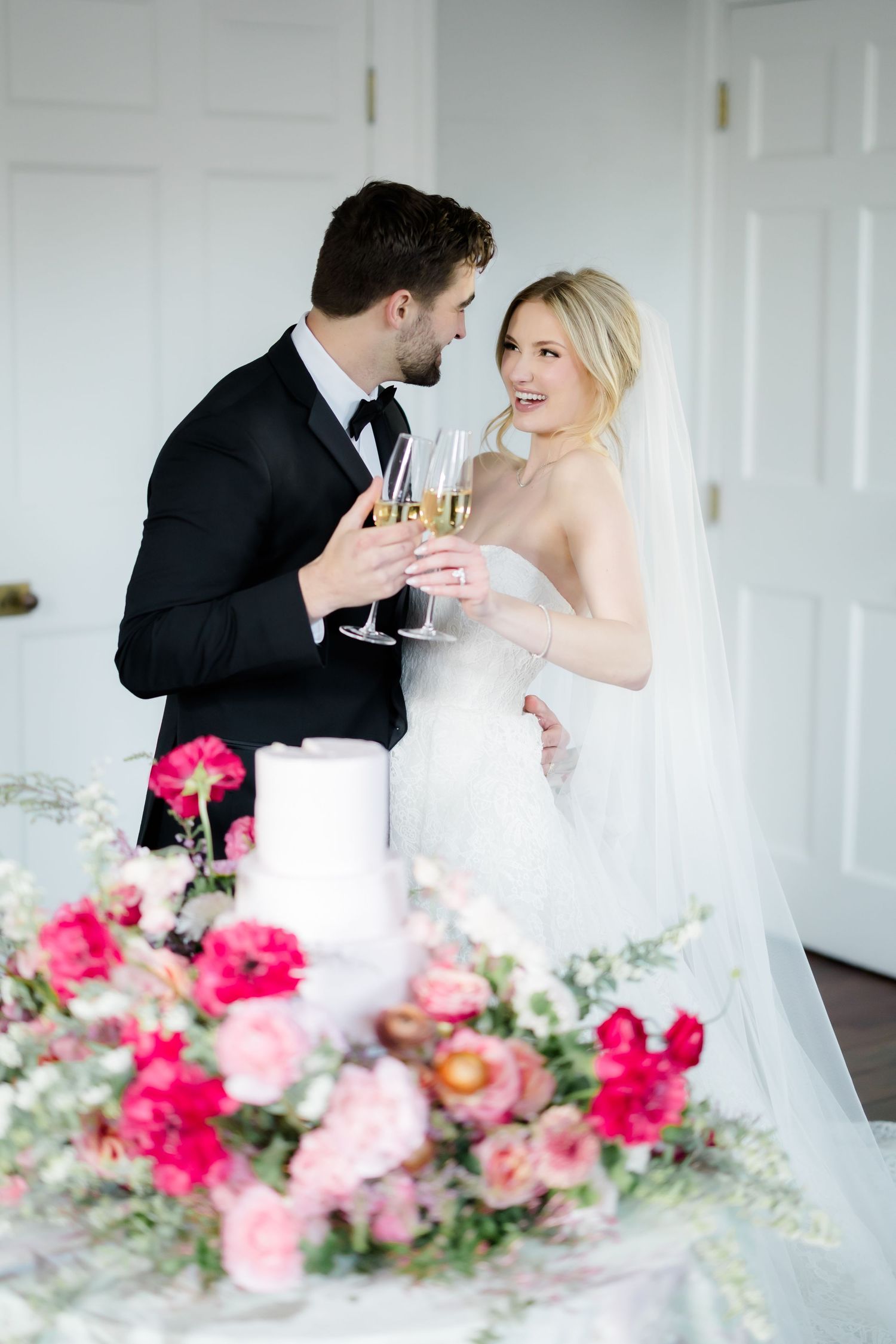Newlyweds share a champagne toast beside a floral wedding cake display with pink and red blooms.