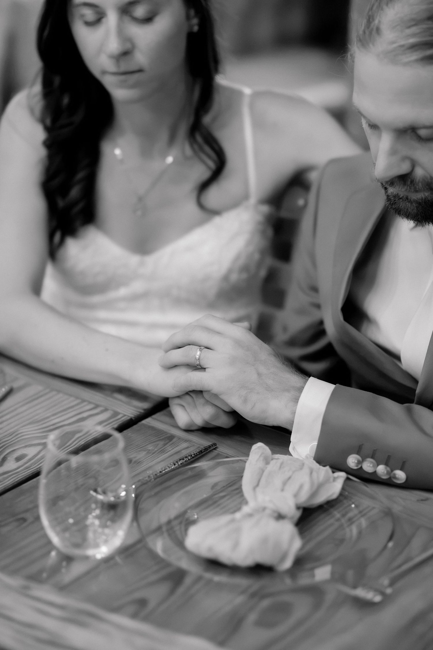 Black and white photo of bride and groom holding hands at wedding reception table with wine glasses.