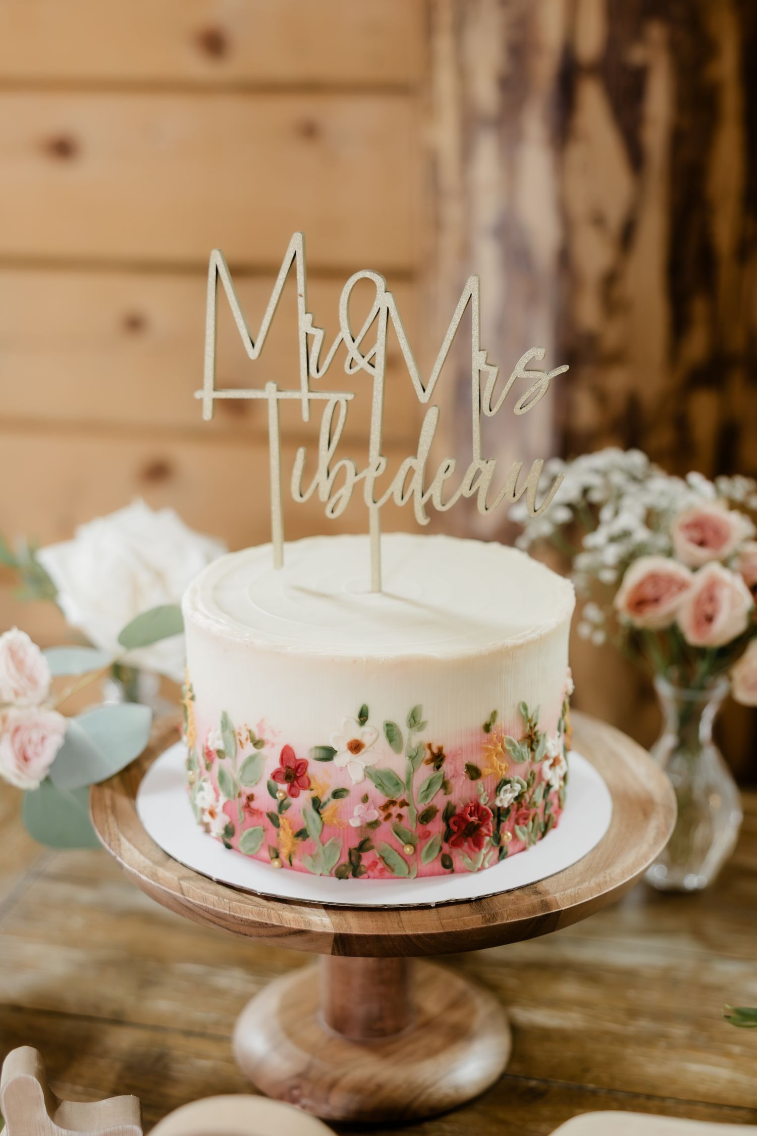 White wedding cake with floral design on wooden stand, topped with Mr & Mrs Tibedeau laser cut cake topper.