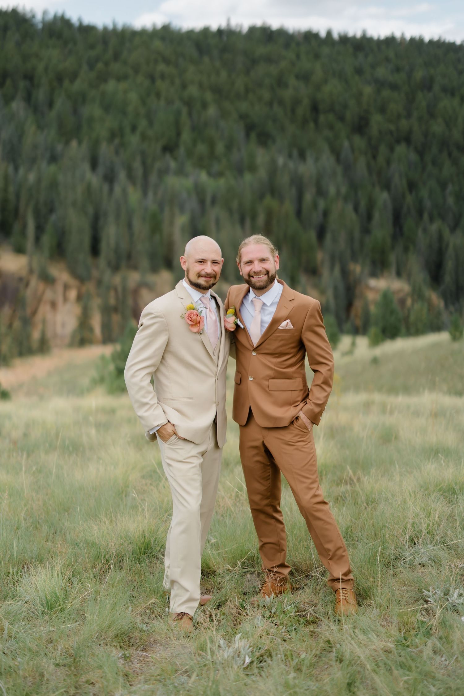 Two grooms in tan and brown suits pose together in a mountain meadow with pine forest backdrop on their wedding day.
