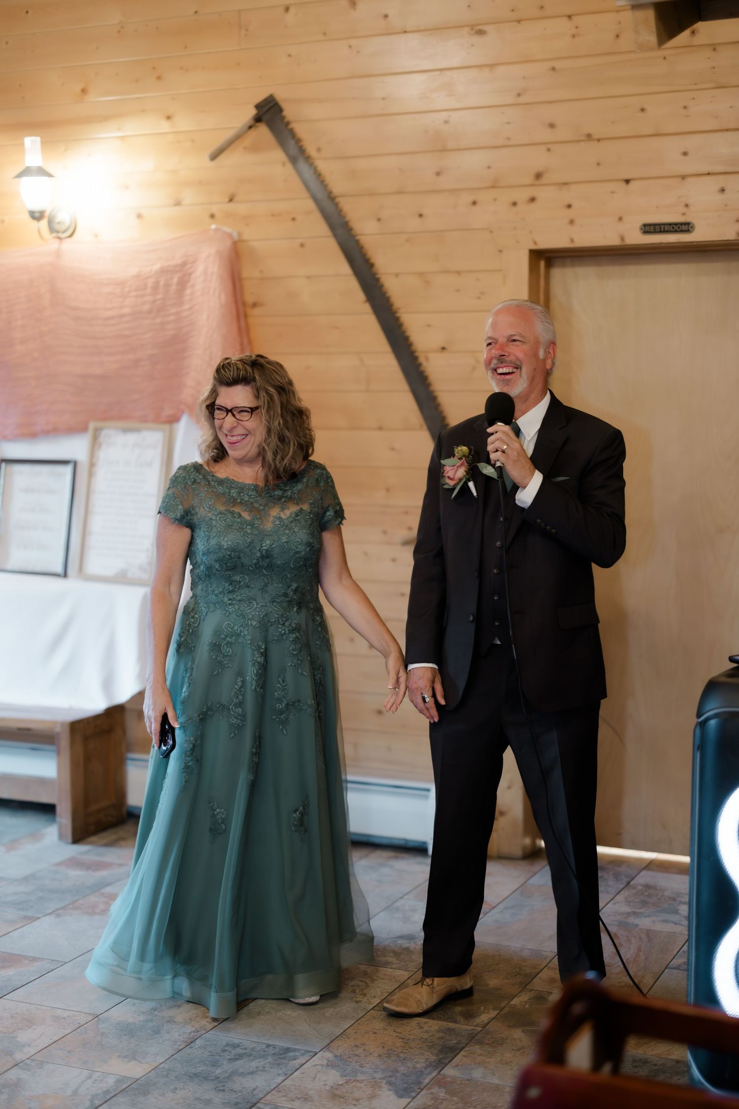 Couple in formal attire smiling at wedding reception, man in black suit holding microphone, woman in green lace gown.