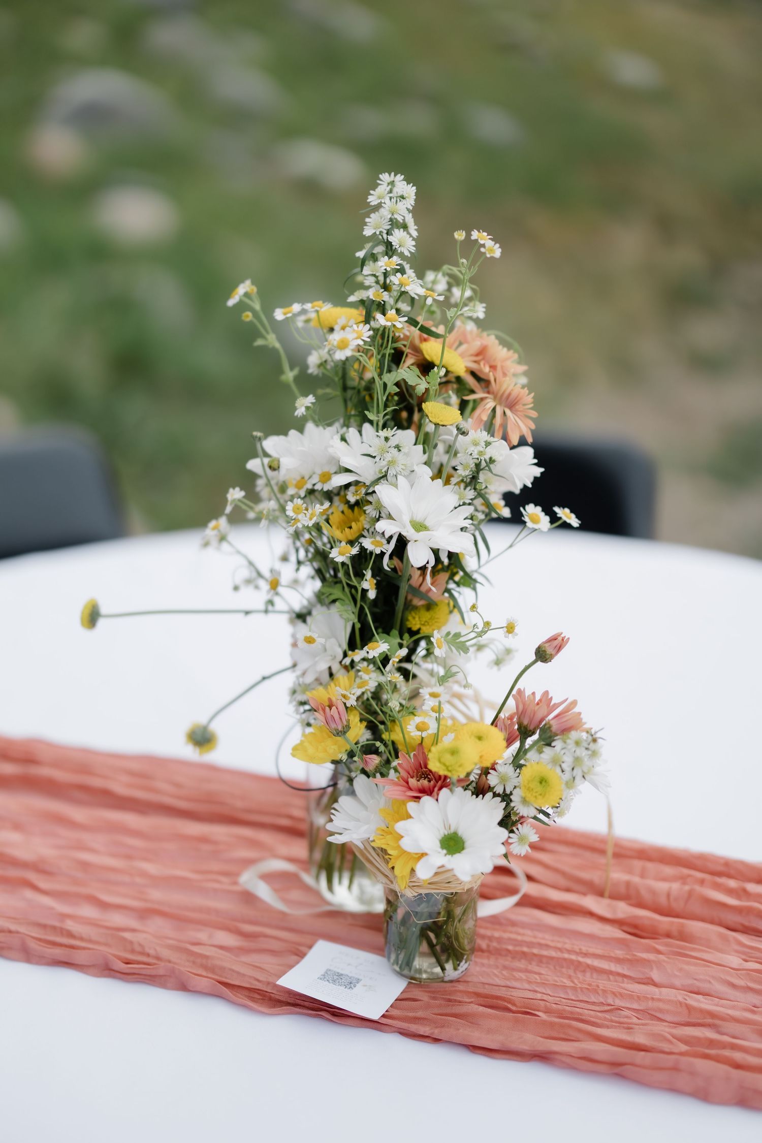 Rustic wildflower centerpiece with daisies and yellow blooms in glass vases on a coral table runner.