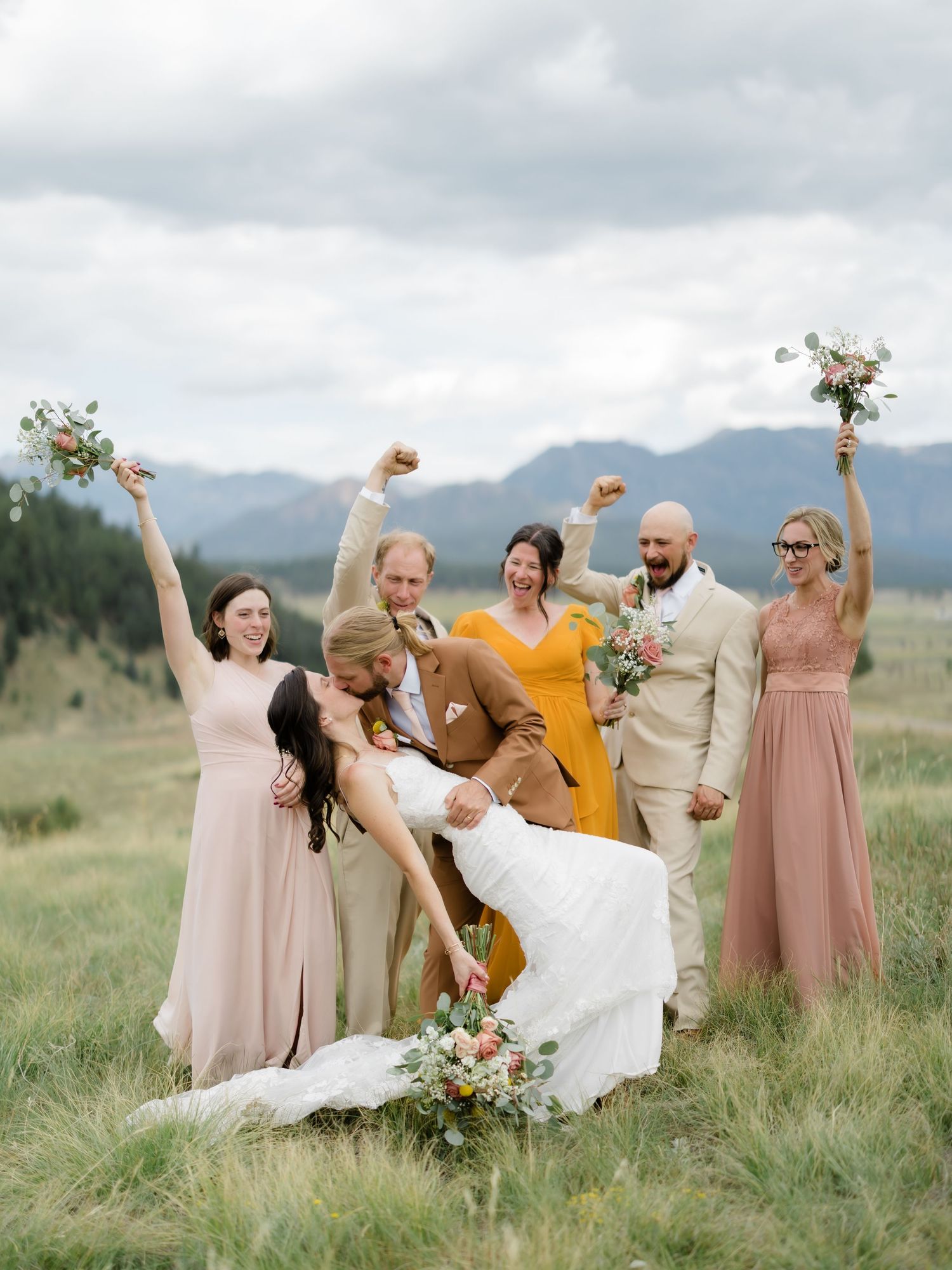 Bride and groom share a dip kiss surrounded by cheering wedding party in blush and mustard gowns with mountain backdrop.
