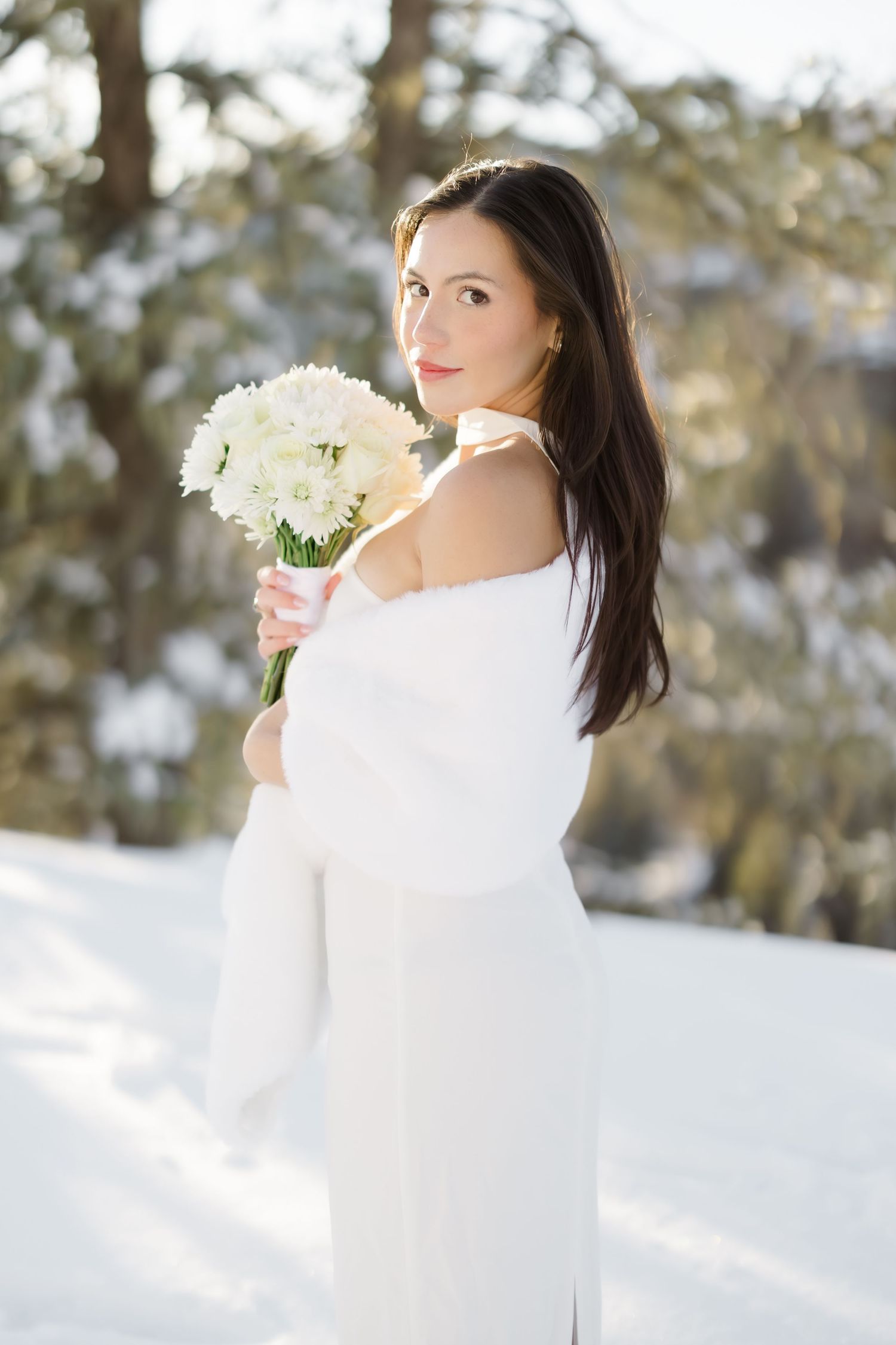 Elegant bride in white dress and fur shawl holding white flower bouquet in a sunny snowy landscape.
