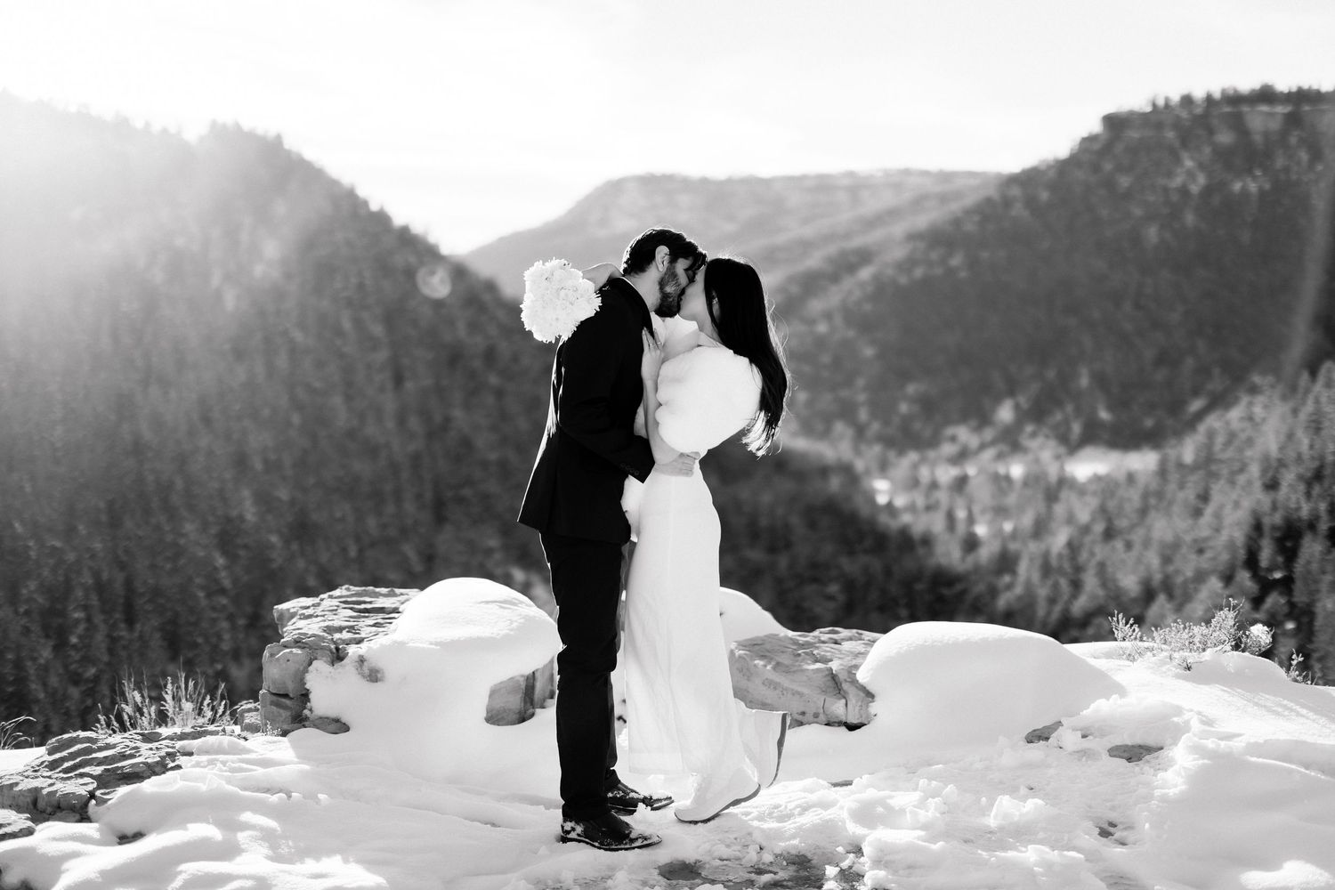 Bride and groom share a romantic kiss on a snowy mountain overlook in a stunning black and white wedding photo.
