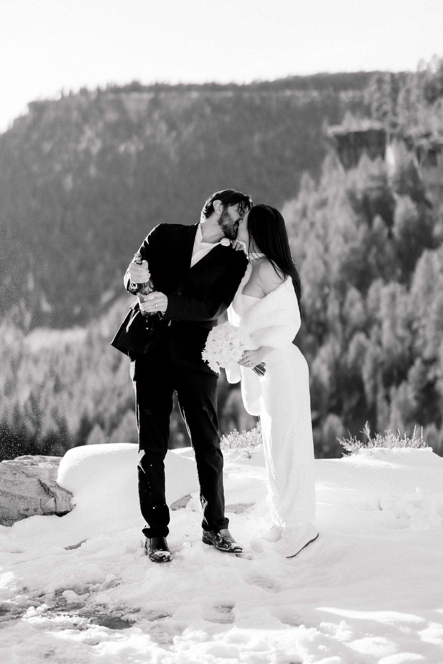 Black and white photo of bride and groom kissing while popping champagne on snowy mountain.