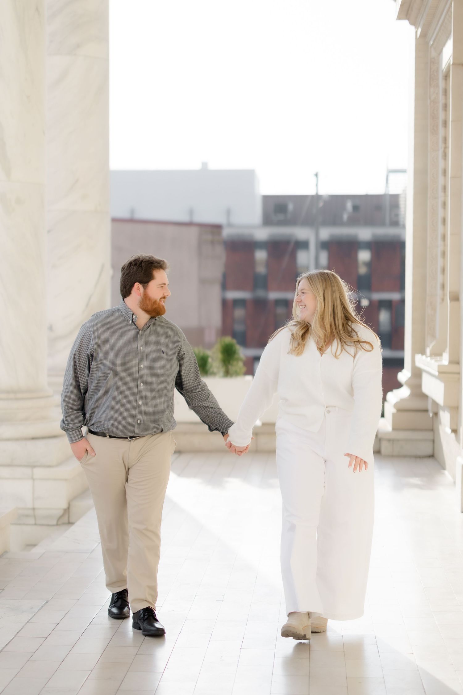 Engaged couple holding hands and walking between grand marble columns, smiling at each other.