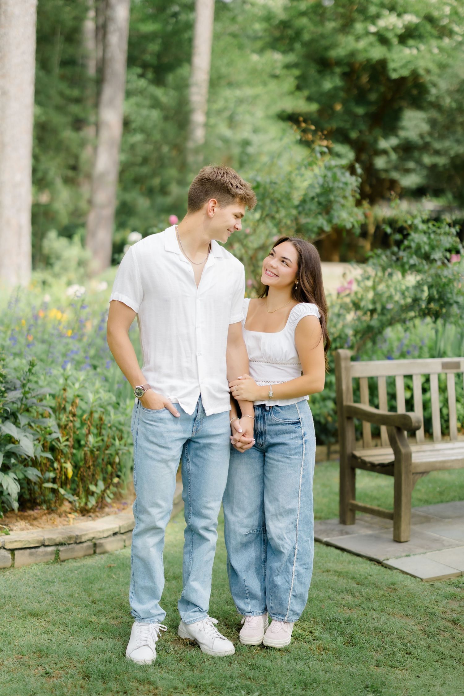 Young couple in matching white tops and jeans holding hands and smiling at each other in a garden.