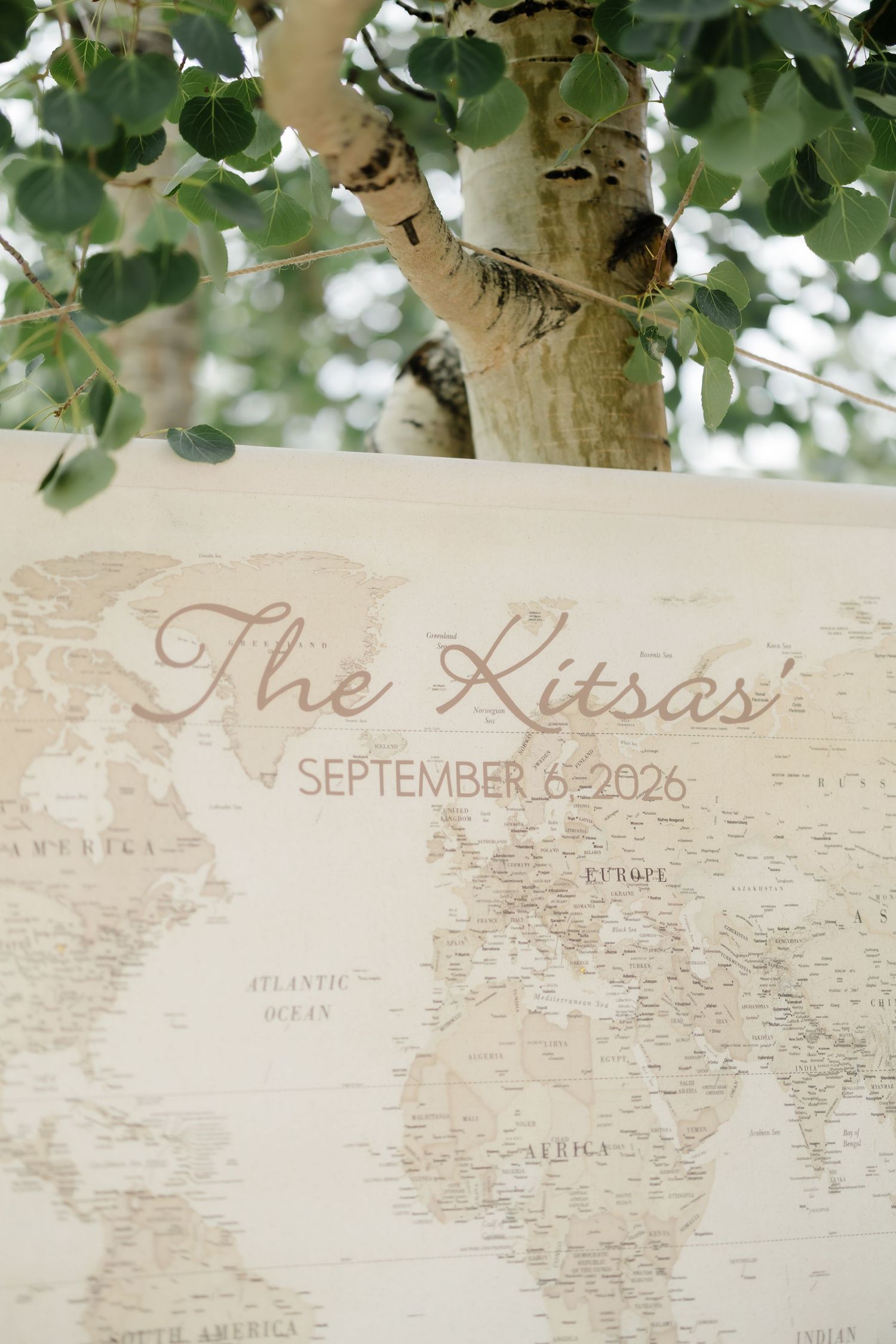 Vintage world map wedding sign for The Kitsas' September 6, 2026, displayed outdoors near a birch tree.