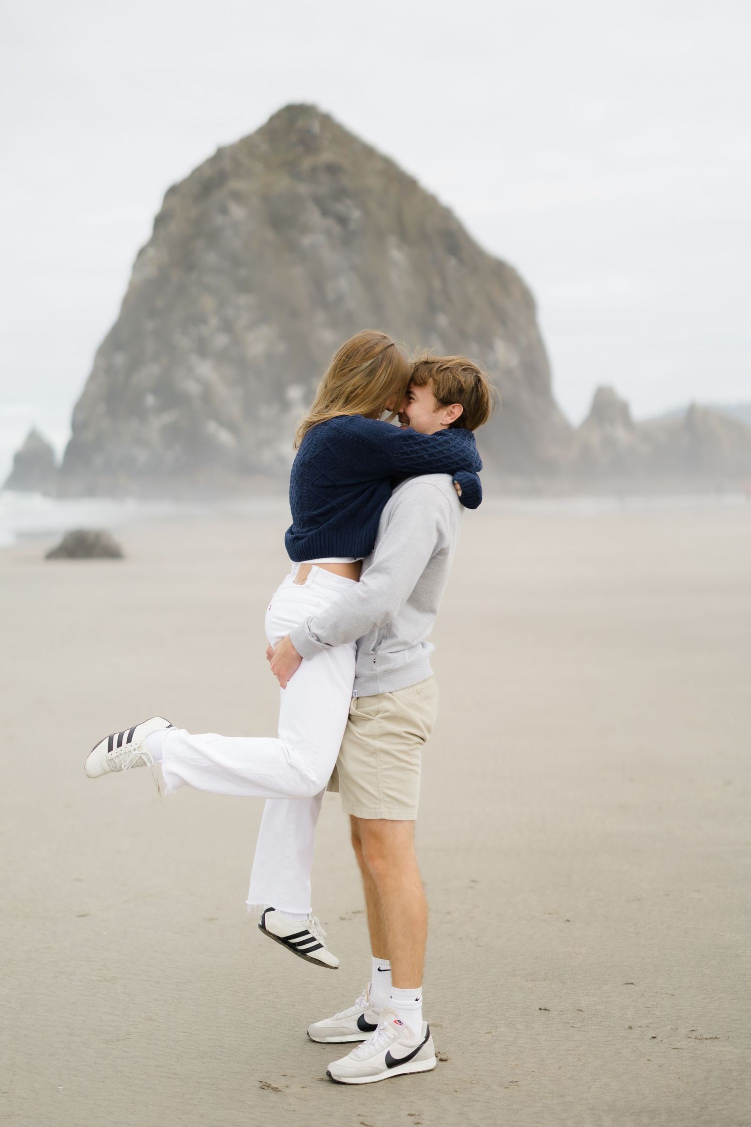 Couple embracing romantically on a misty beach with Haystack Rock in the background at Cannon Beach, Oregon.