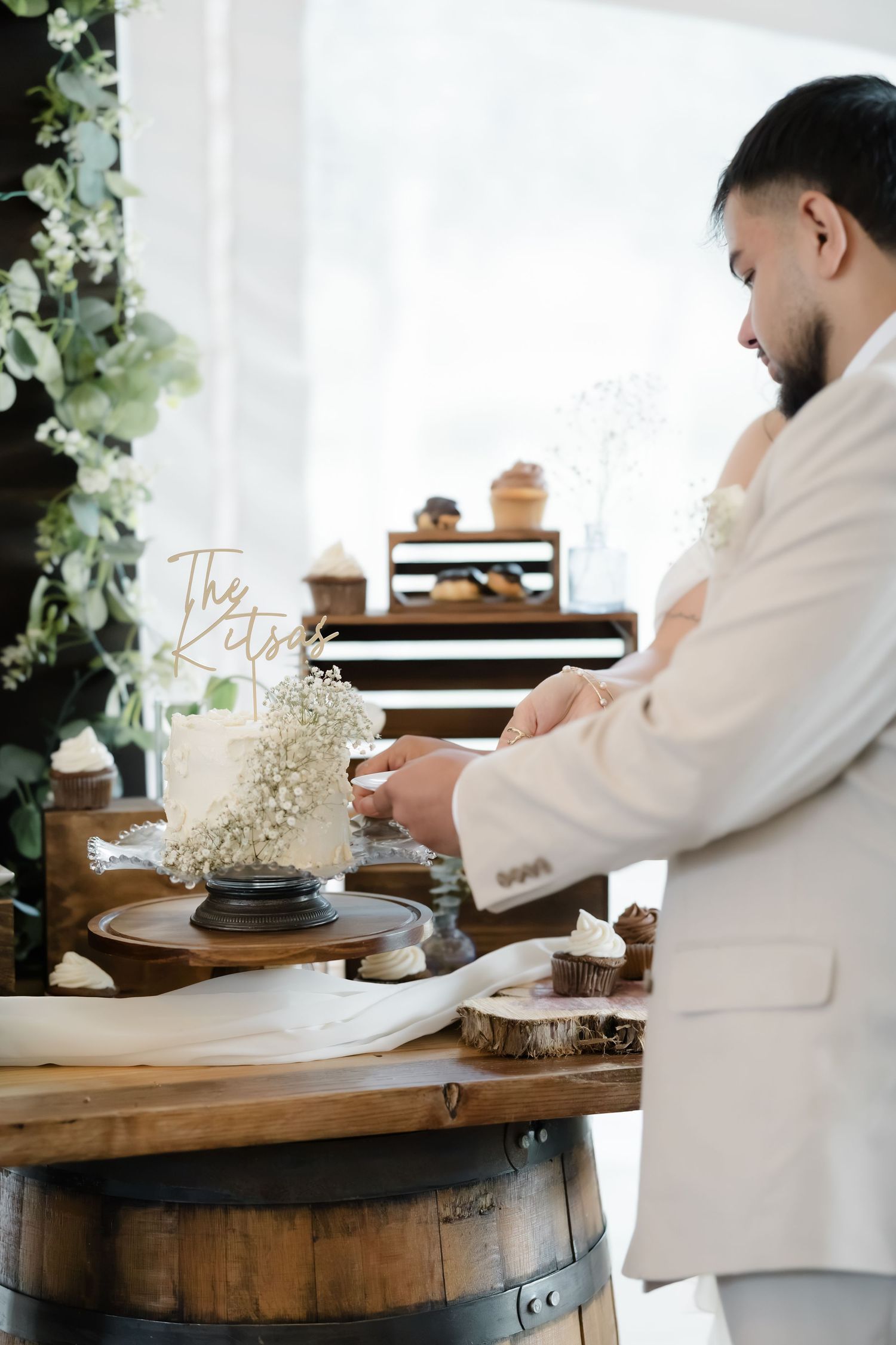 Groom in white suit cutting wedding cake on rustic barrel table with floral decor and baby's breath arrangements.
