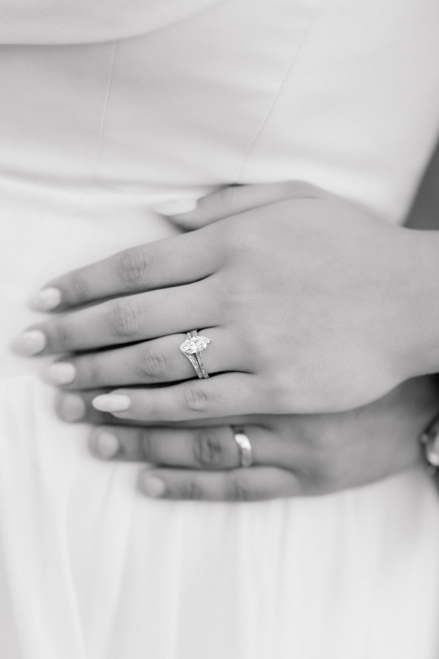Black and white close-up of bride's hand wearing a solitaire diamond engagement ring over white wedding dress.