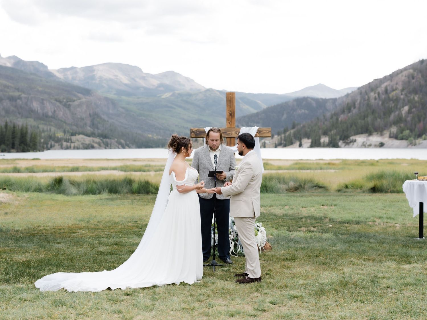 Outdoor mountain wedding ceremony with bride and groom exchanging vows before a rustic cross altar beside a scenic lake.