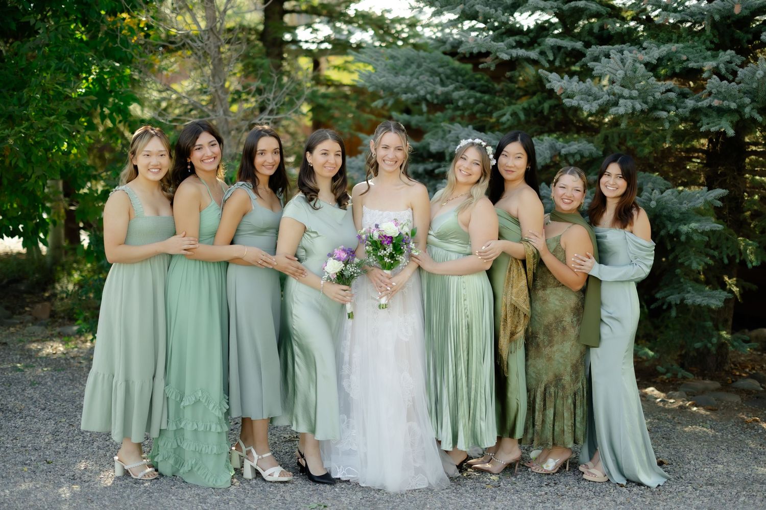 Bride in white gown poses with eight bridesmaids in mismatched sage green dresses, holding bouquet outdoors.