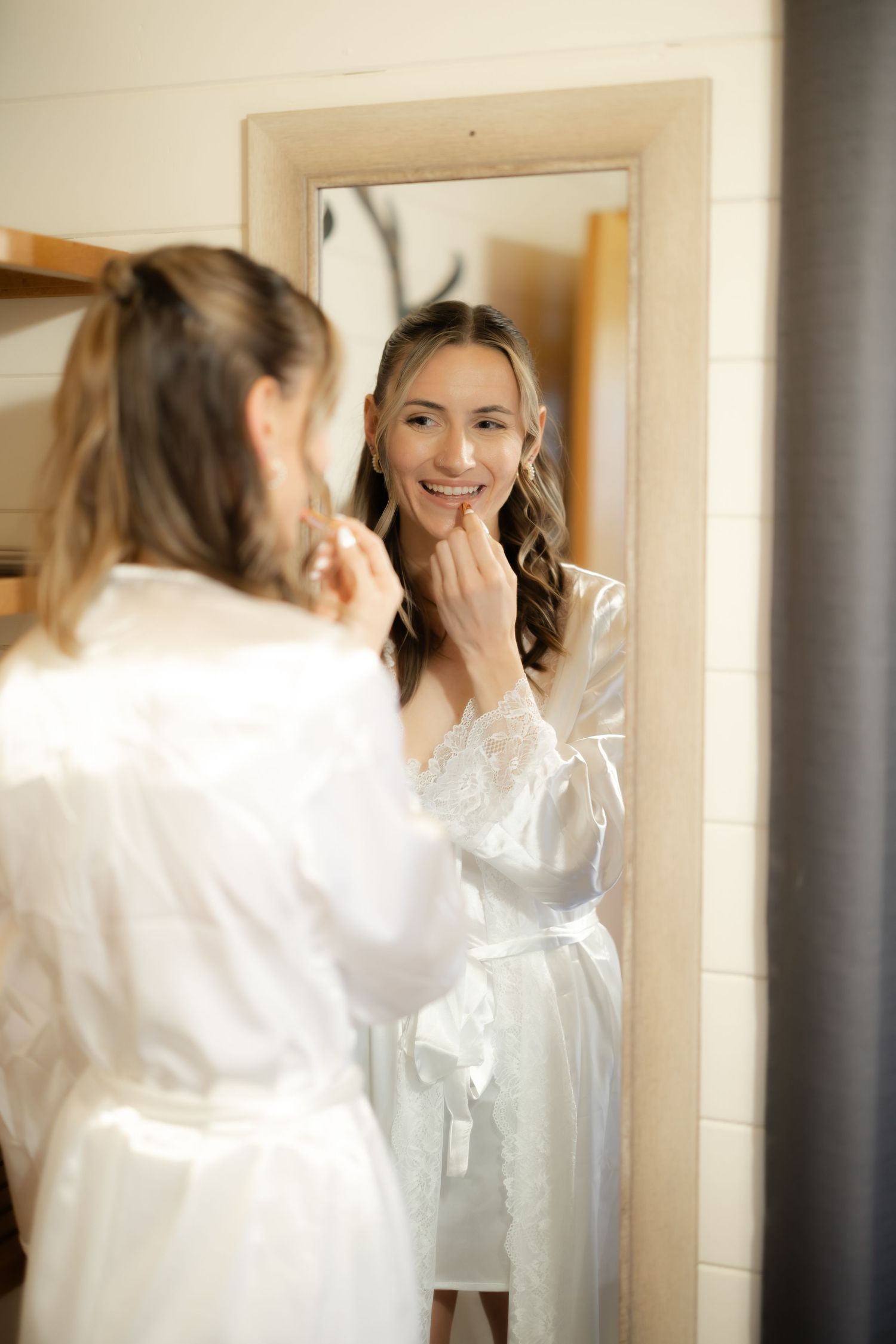 Smiling bride in white lace robe applying makeup while looking in a full-length mirror on her wedding day.