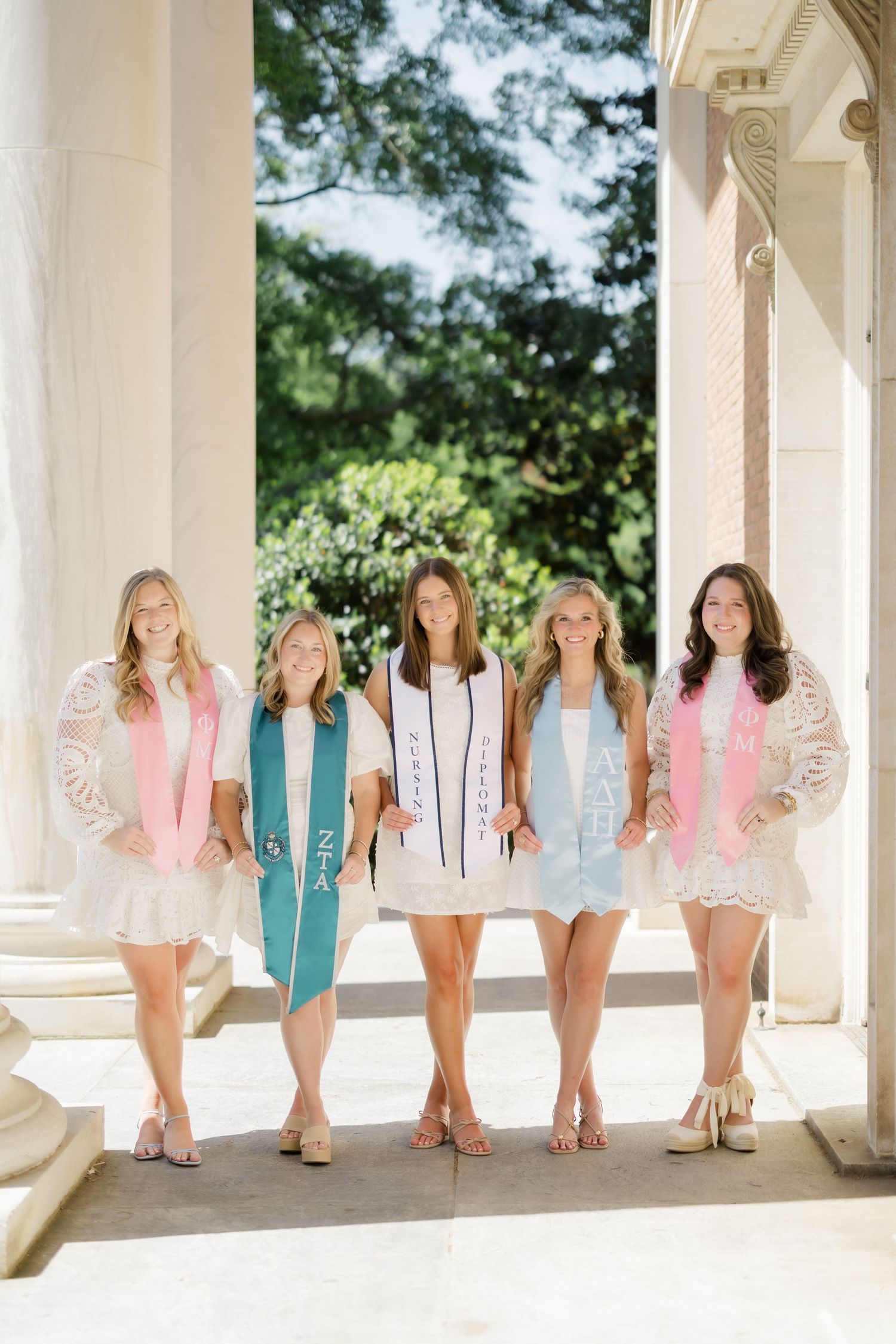 Five college women in white dresses wearing colorful graduation stoles pose between pillars outdoors.