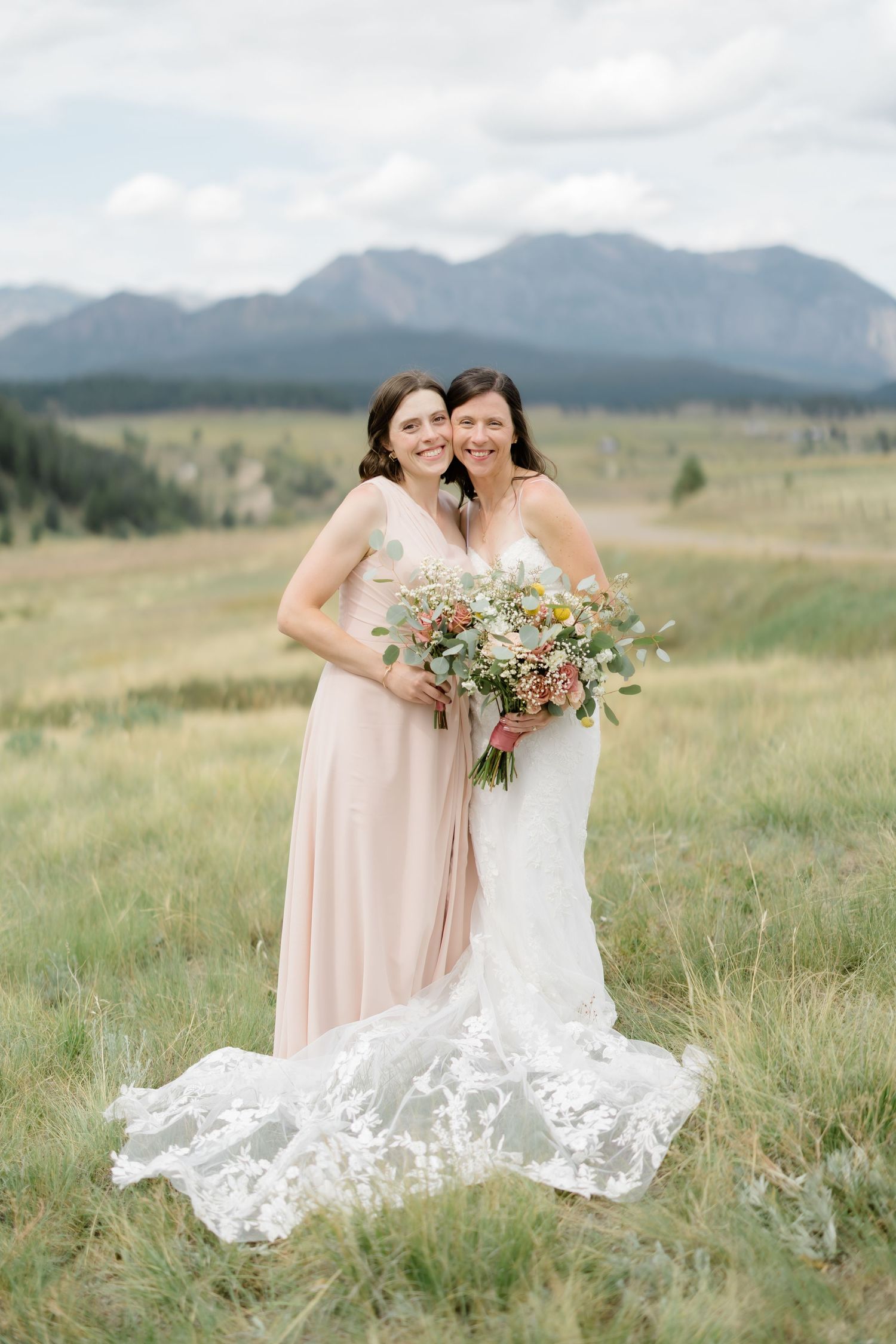 Bride in lace gown and bridesmaid in blush dress pose with bouquet in mountain meadow.