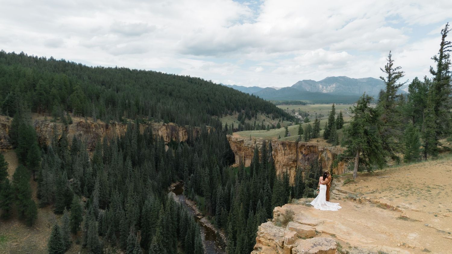 Couple shares romantic wedding moment on rocky cliff edge overlooking lush pine forest and mountain valley landscape.