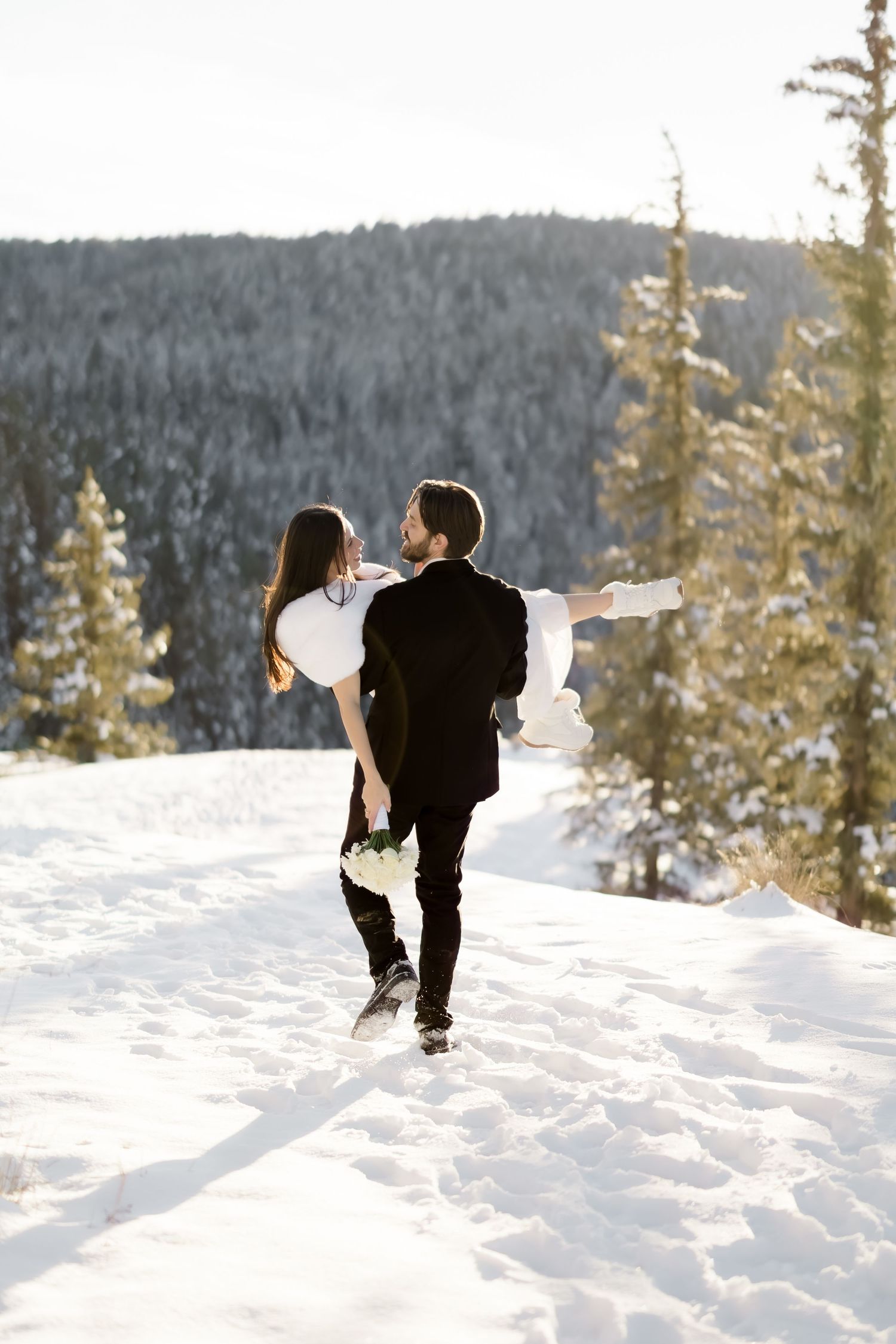 Groom carries bride through snowy mountain landscape surrounded by snow-covered evergreen trees.