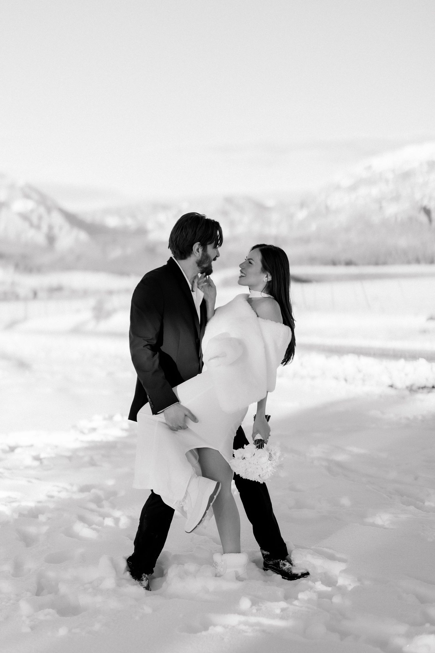 Black and white photo of a groom lifting his bride in a snowy mountain landscape during their winter wedding.