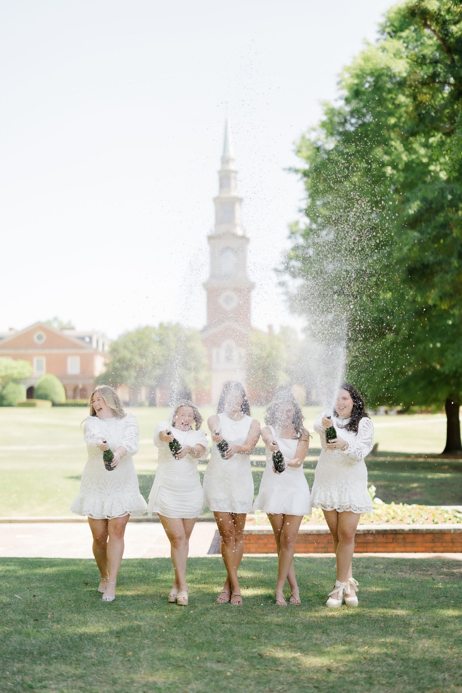 Five women in white dresses celebrate outdoors by popping champagne bottles near a brick church steeple.