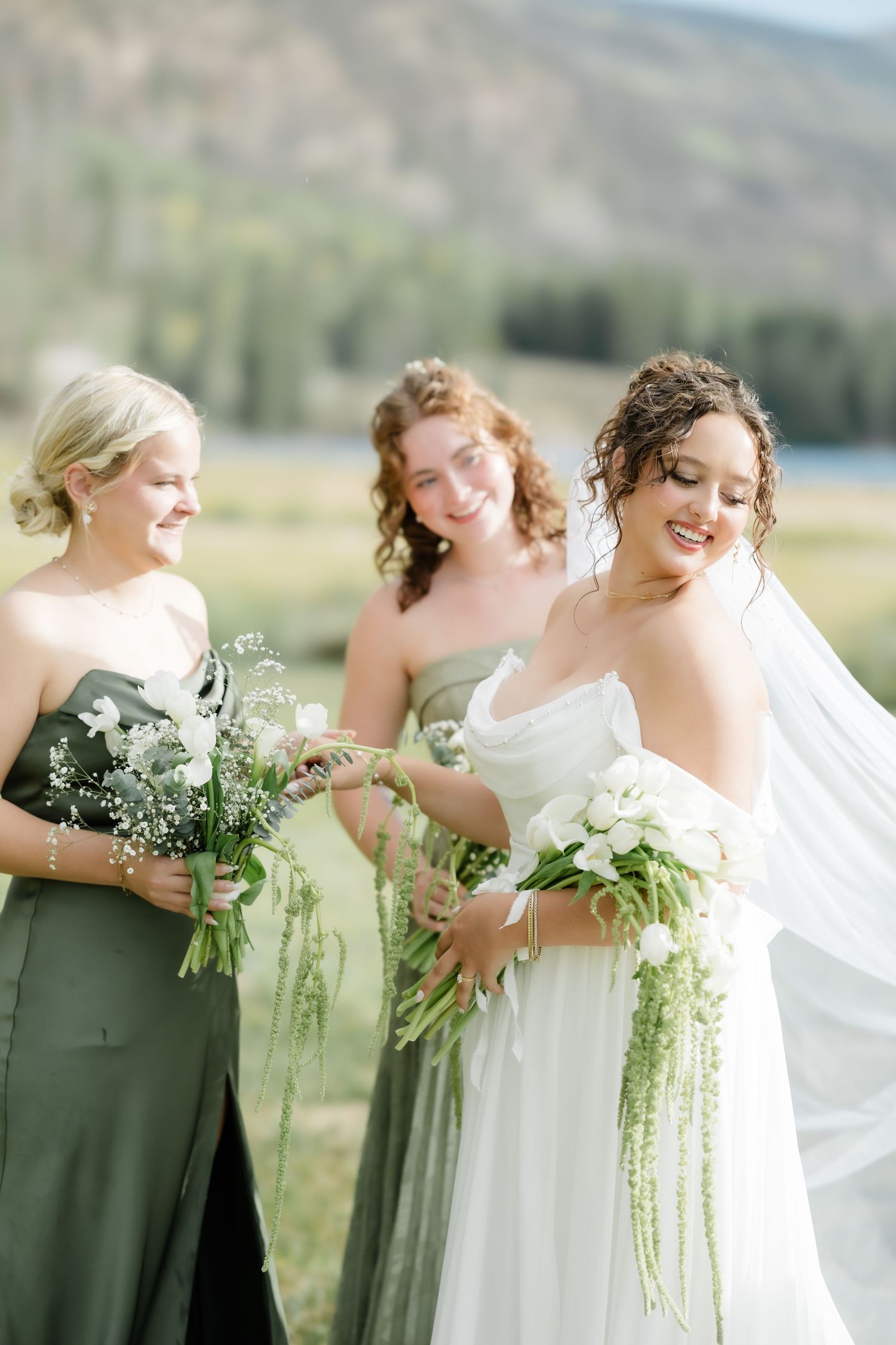 Bride in white gown with bridesmaids in sage green dresses holding white floral bouquets outdoors.