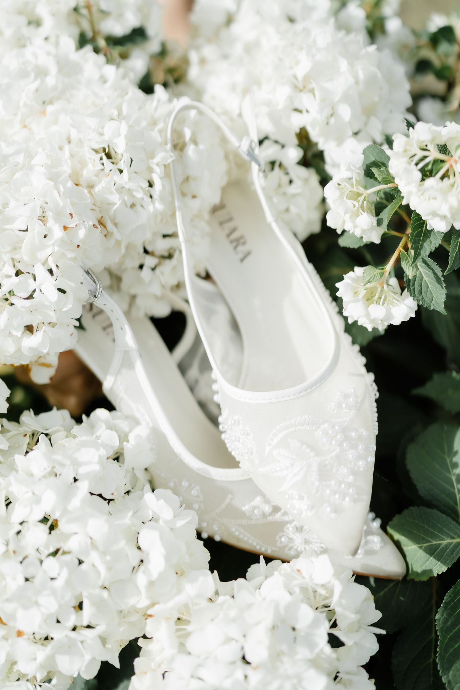 White lace bridal heels with pearl embellishments resting among white hydrangea flowers.