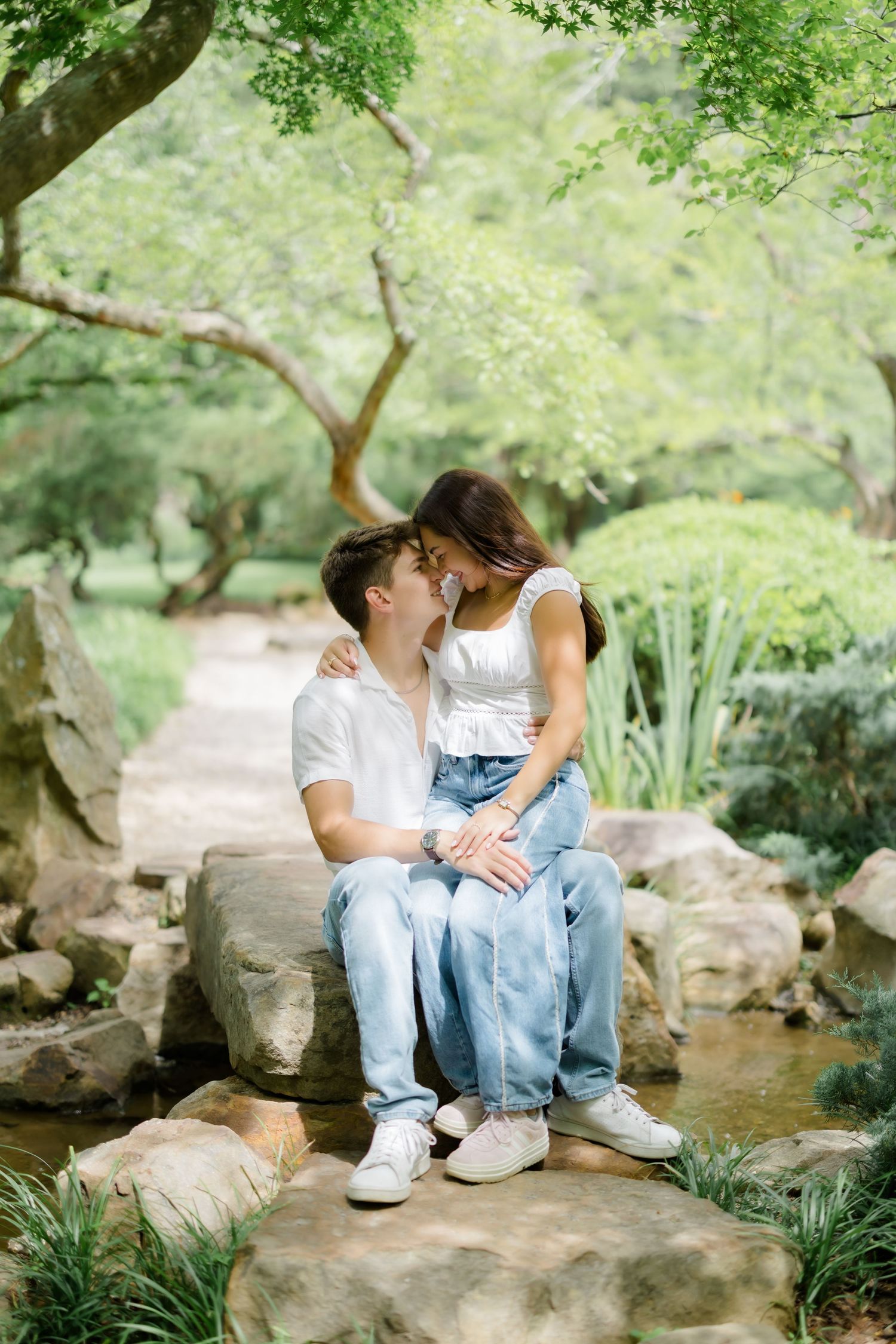Couple sitting on rocks in a lush green park, leaning in for a kiss, wearing white tops and jeans.