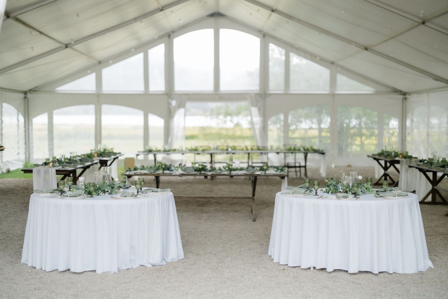 Elegant wedding reception tent with white draped tables, greenery garlands, and large windows overlooking a scenic outdoor view.