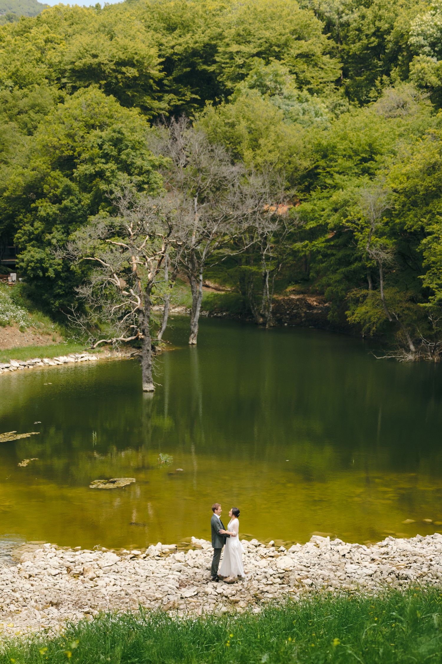 A couple stands by a serene mountain lake surrounded by lush green forest and rocky shoreline.