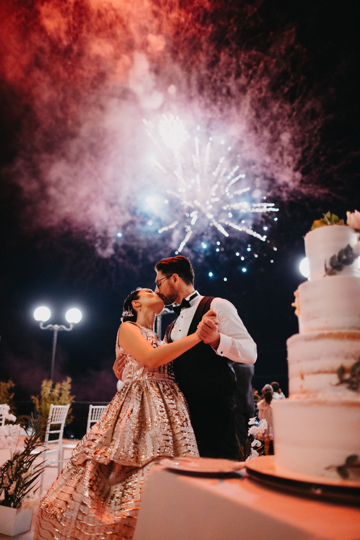 A wedding couple dances under dramatic fireworks and a starlit sky next to their elegant white wedding cake.