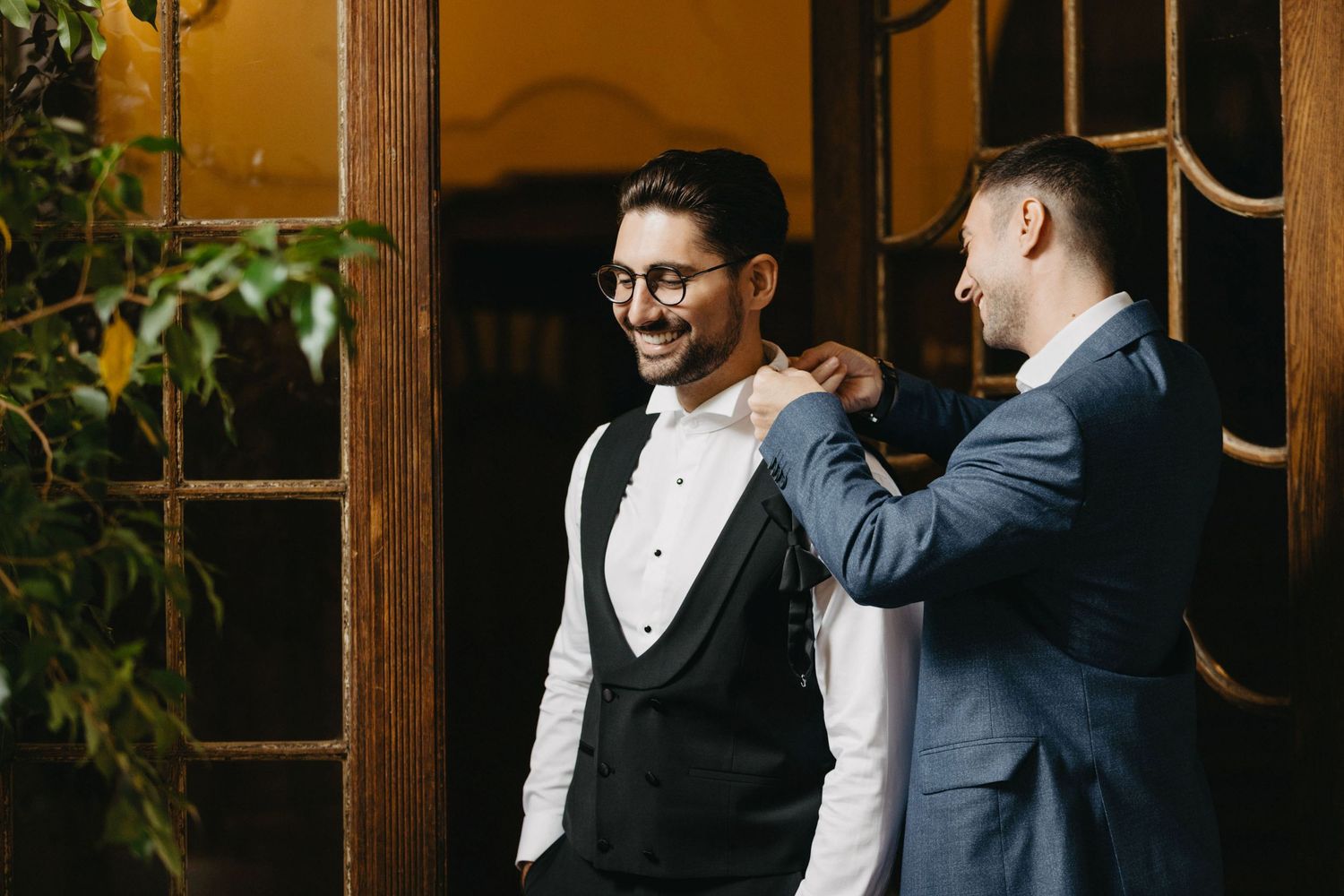 Two well-dressed men in formal attire sharing a candid moment by a wooden doorway.
