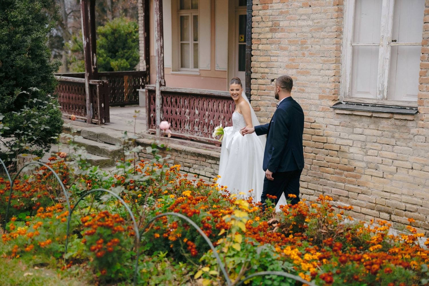 A couple in wedding attire stands near vibrant orange flowers outside a historic brick building.