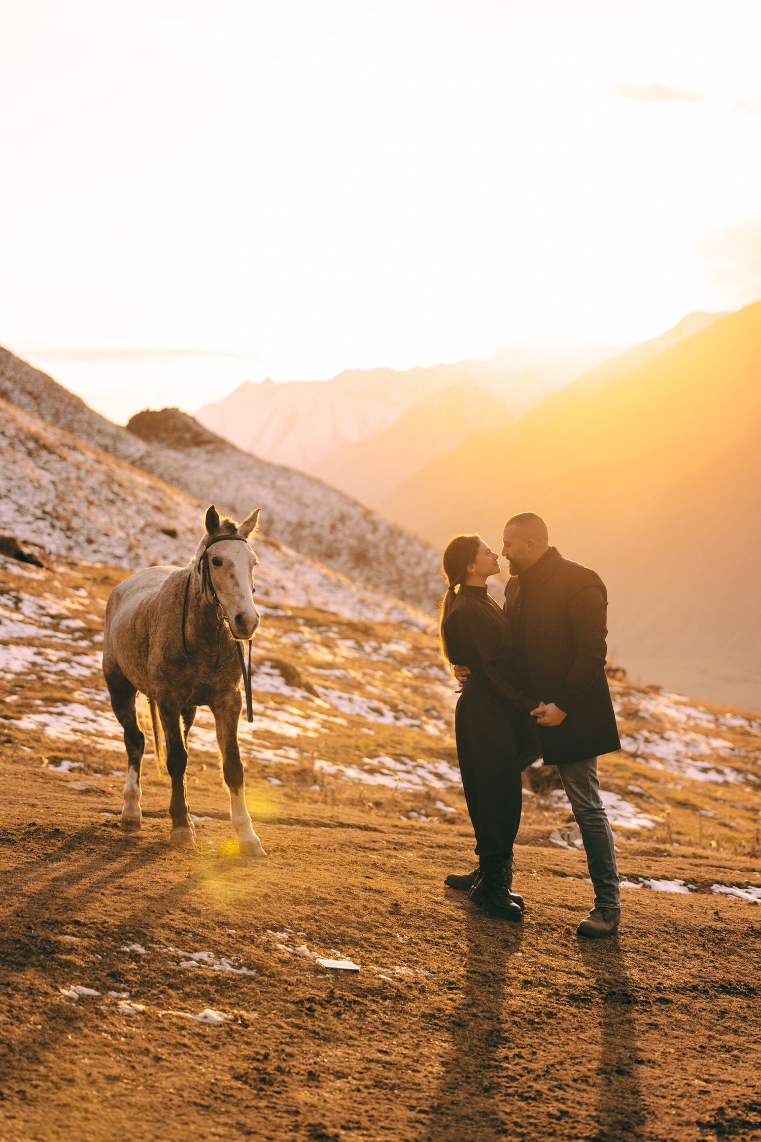 Couple kneeling in snow with horse standing nearby during golden hour in mountains.