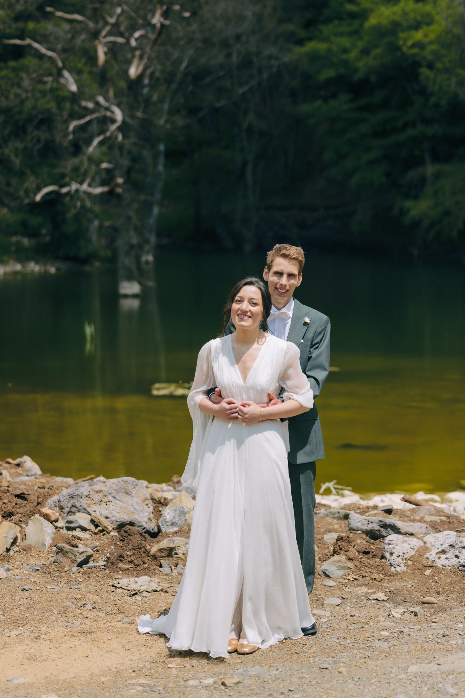 A romantic wedding photo by a serene lake with mountains in the background.