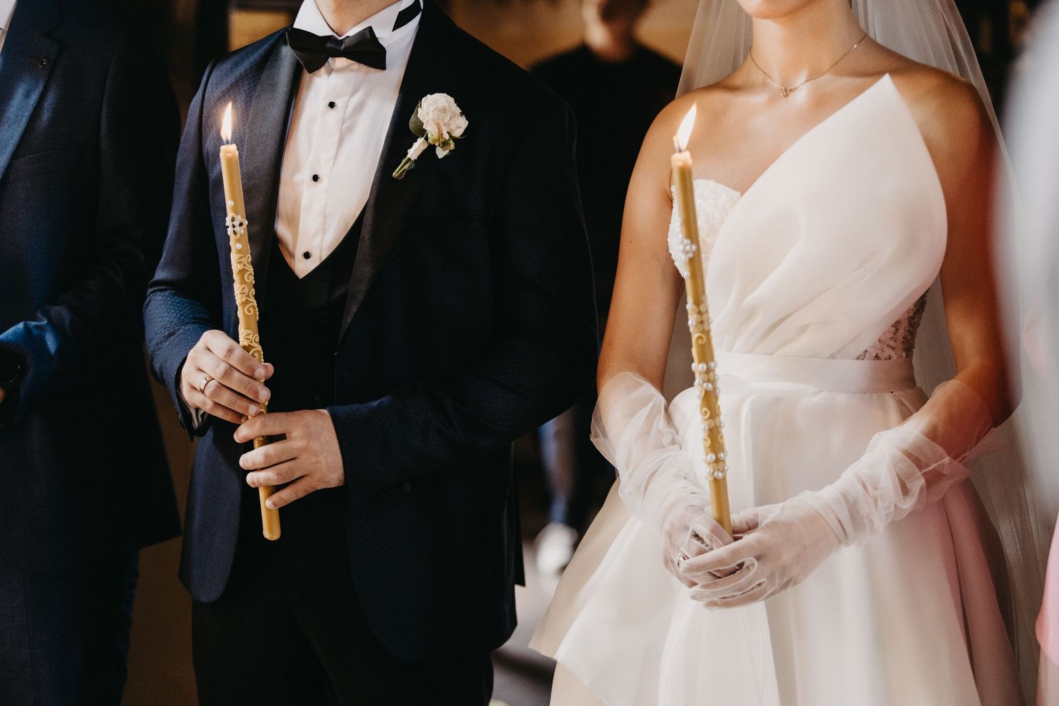 Wedding party members hold lit ceremonial candles in formal attire.