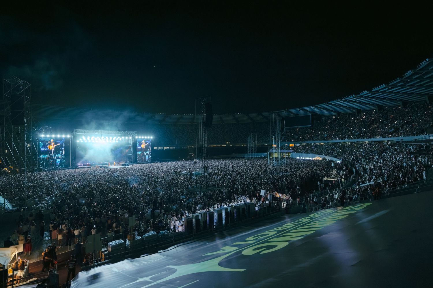 Massive stadium concert with bright stage lights illuminating packed audience.