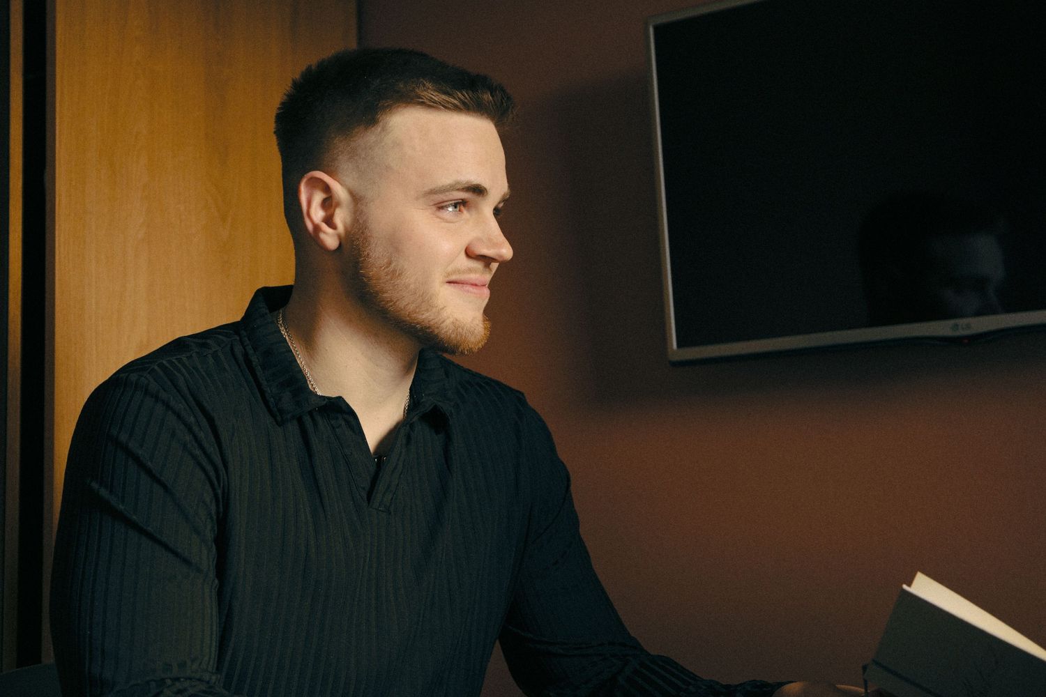 Person in black attire sits at a wooden table in a dimly lit room during an intimate moment.