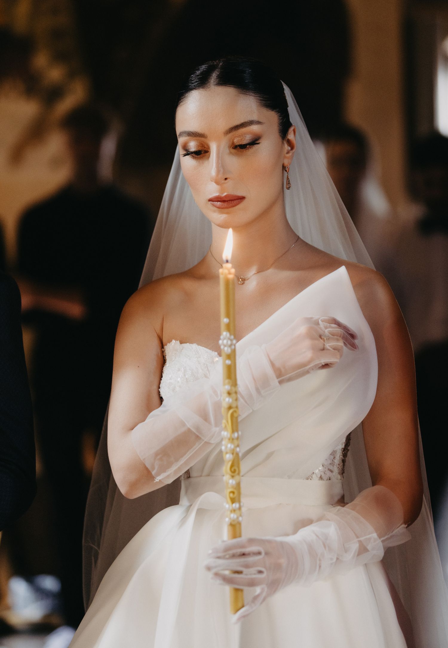 Bride in white gown holds decorative candle during traditional wedding ceremony.