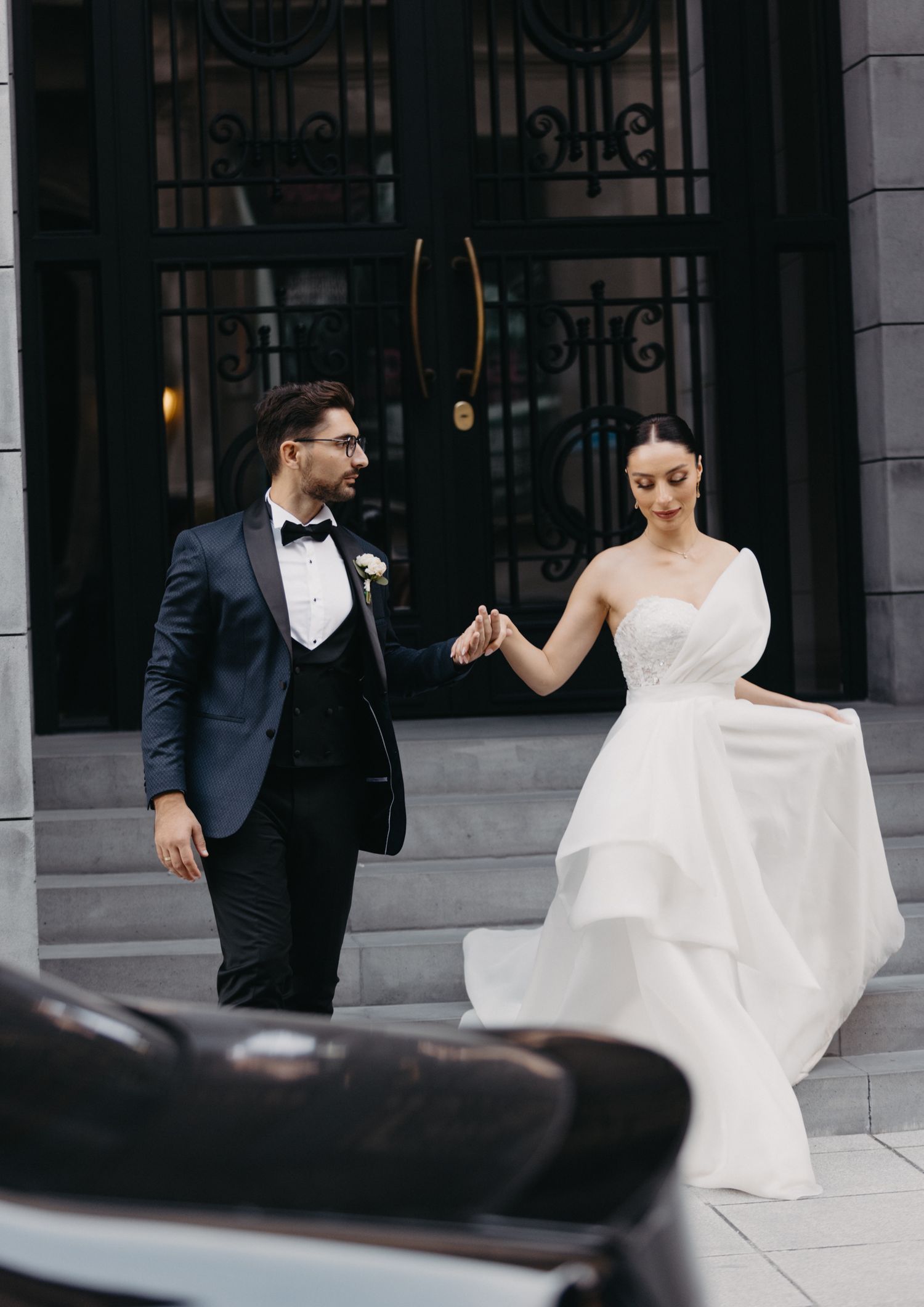 Couple in formal wedding attire descends elegant stone steps with ornate iron gates.