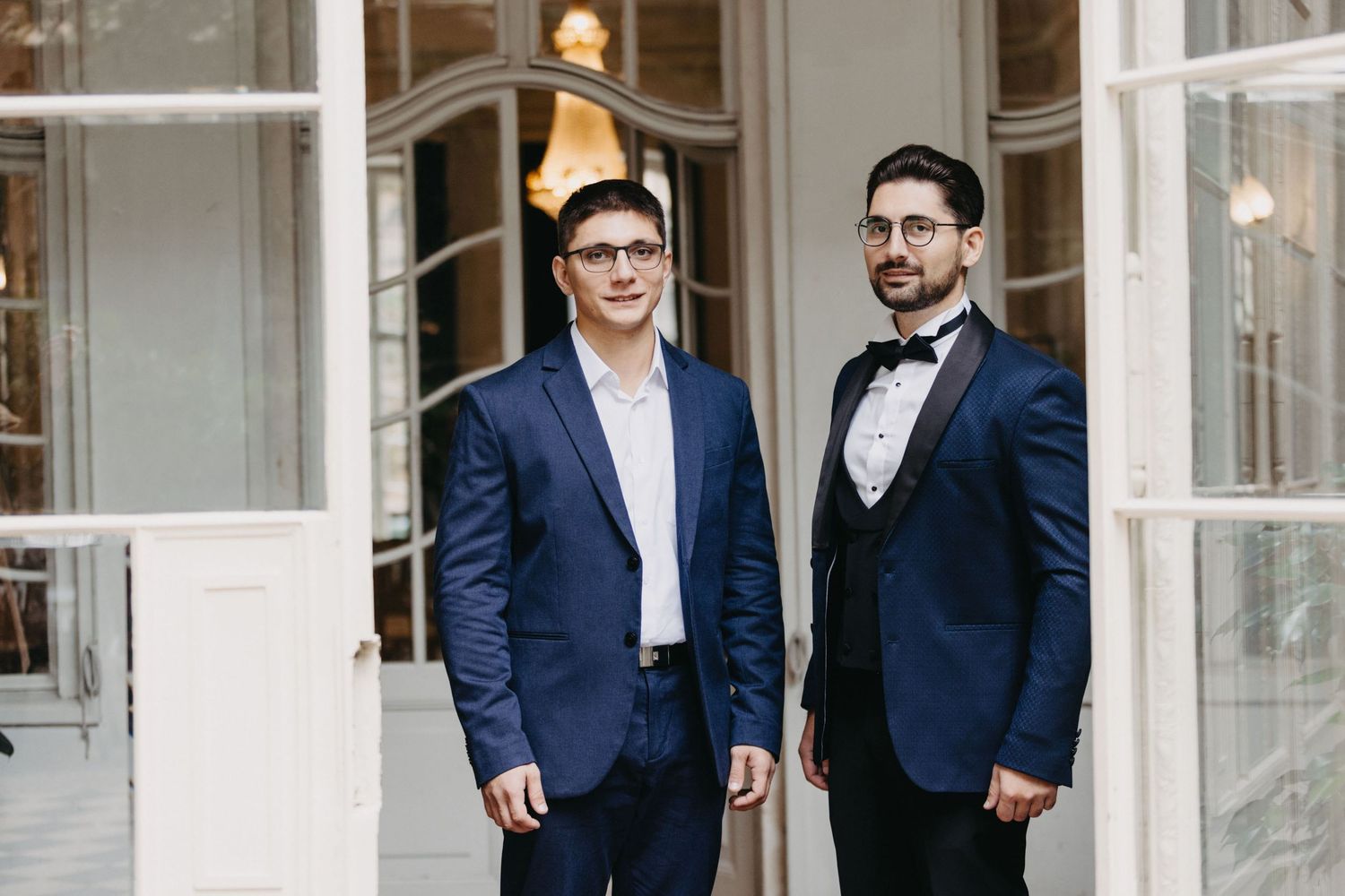 Two men in formal suits posing together in front of elegant white French doors.