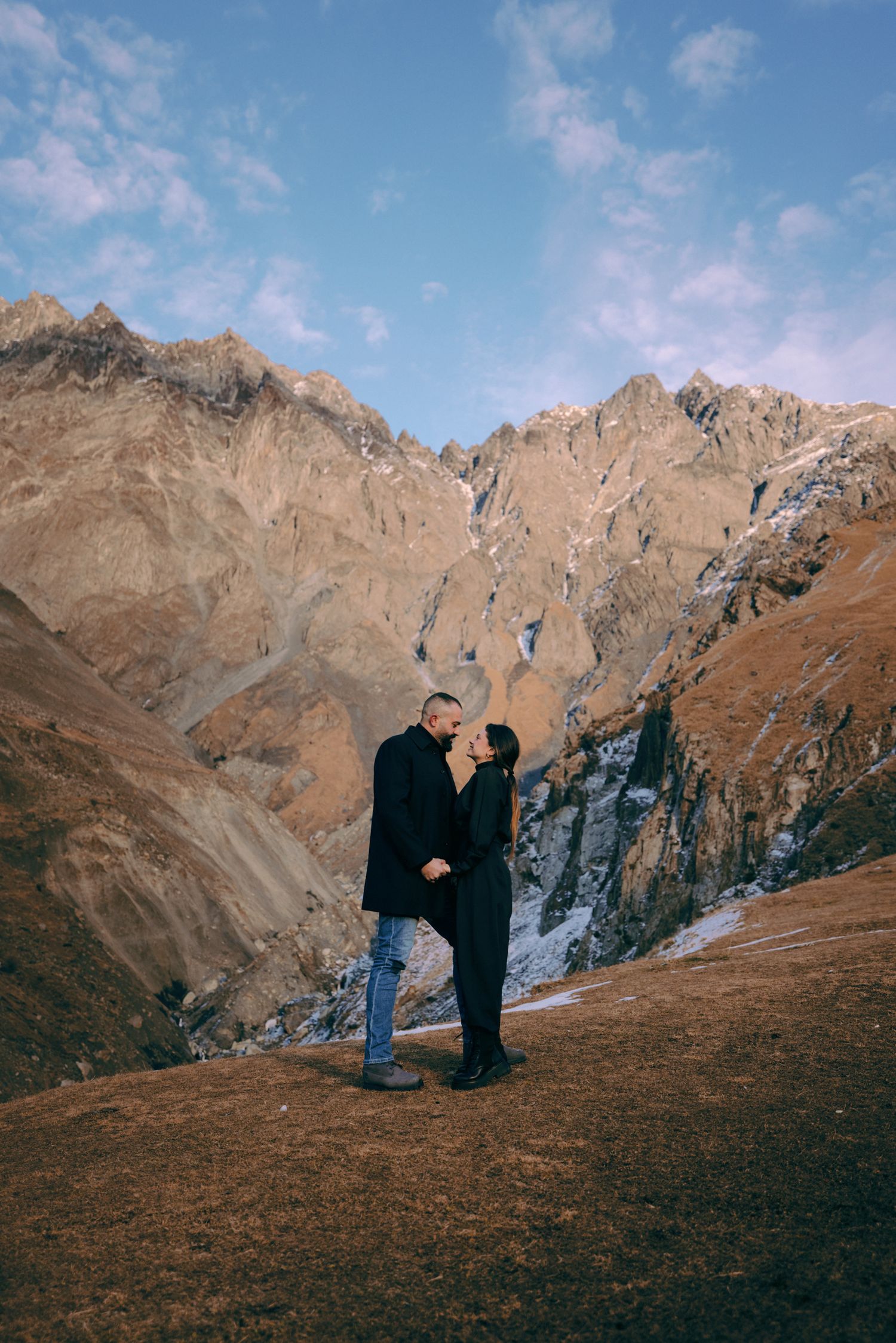 Two people stand against dramatic rocky mountain peaks under blue skies.