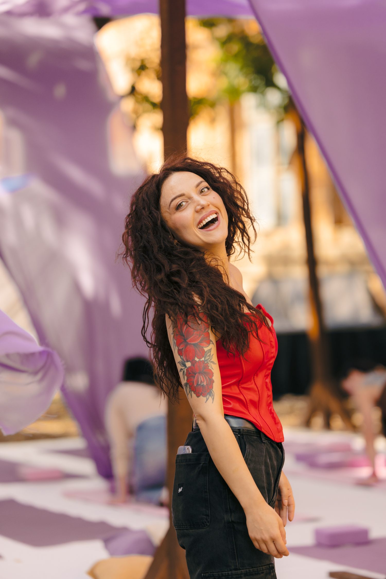 Woman in red top laughing joyfully against purple fabric backdrop at outdoor event.