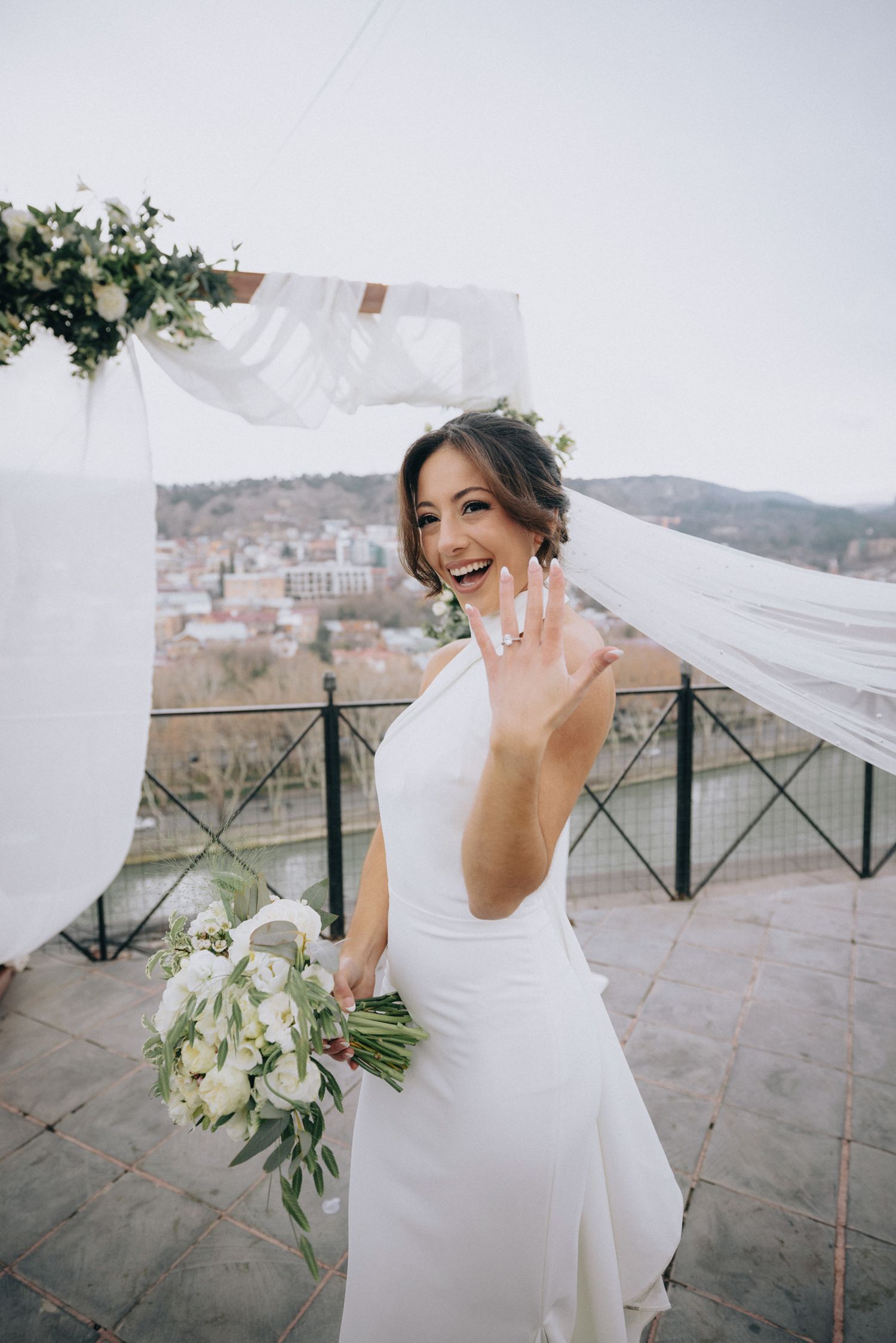Bride in elegant white halter dress holds bouquet while posing on scenic overlook terrace.