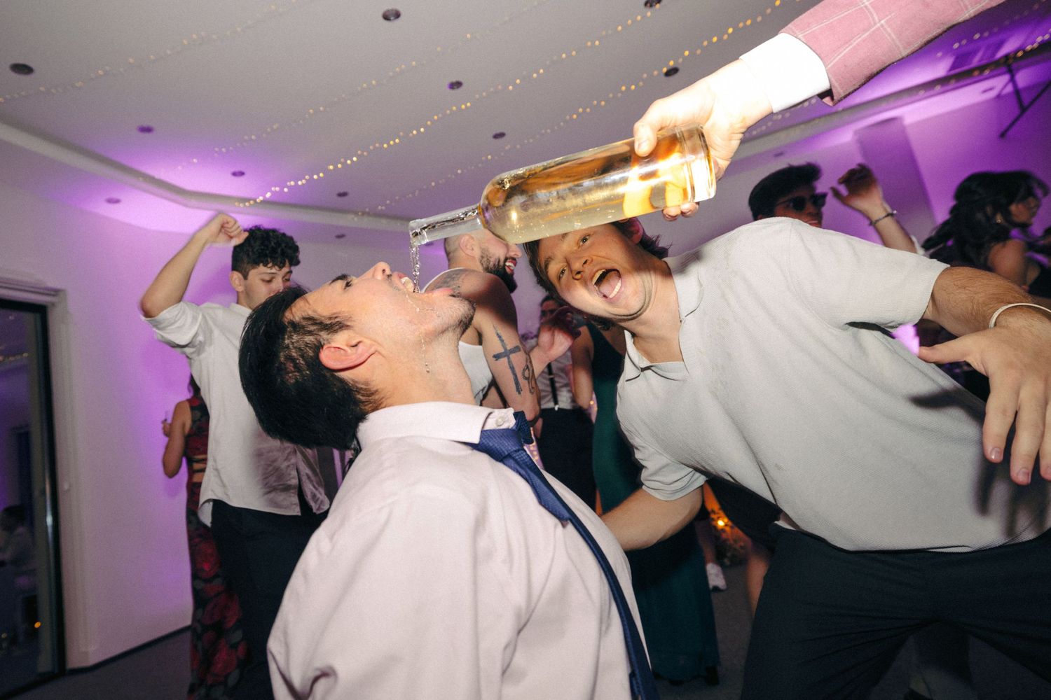 Wedding reception party scene with dancing guests under pink and purple lighting.